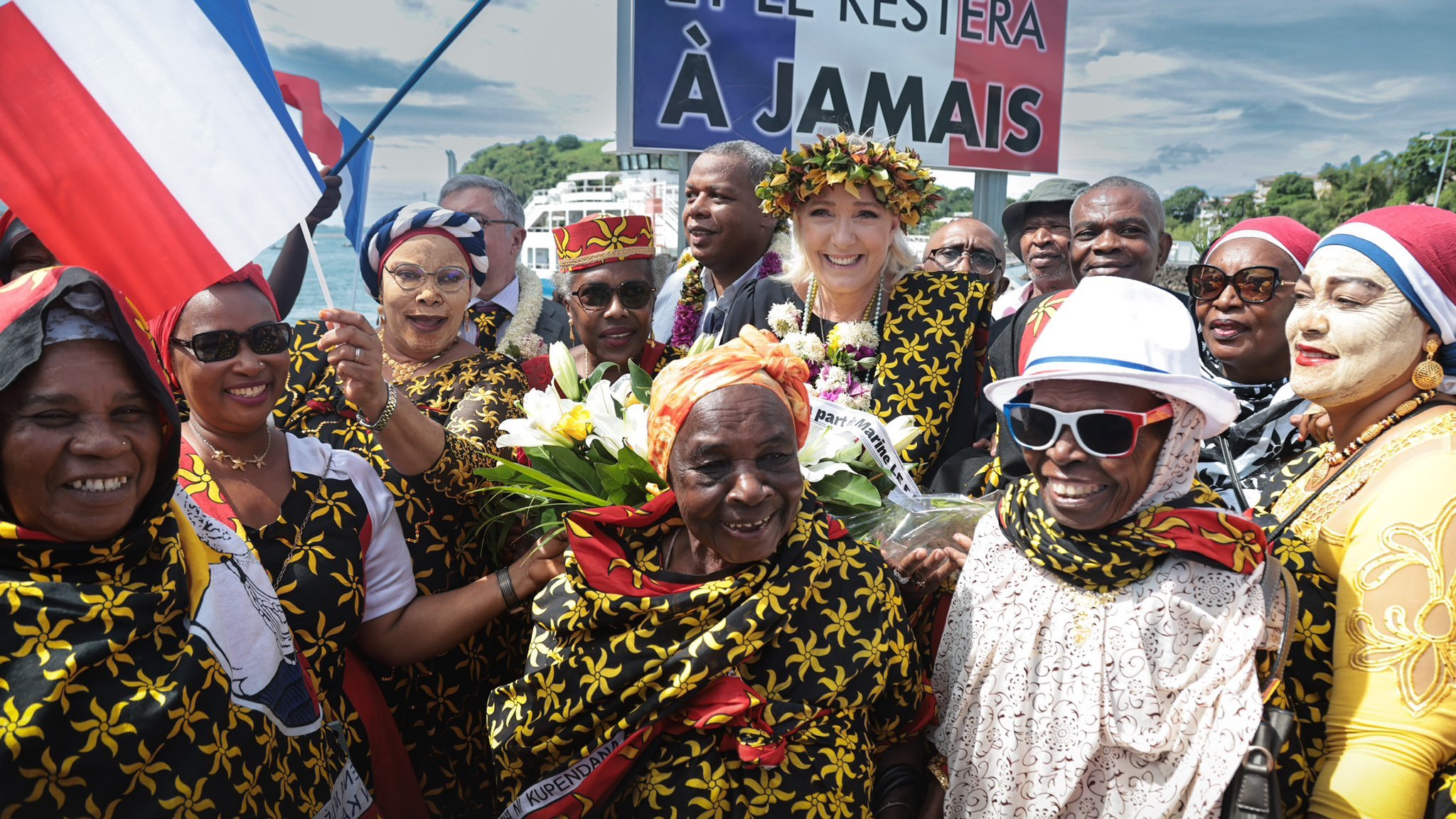 L'image représente une scène festive, probablement lors d'une célébration culturelle ou d'un événement politique. Au centre, plusieurs femmes et hommes portent des vêtements colorés, souvent ornés de motifs floraux jaunes sur fond noir. Certains participants tiennent des bouquets de fleurs tandis que d'autres brandissent des drapeaux français. Au fond, un grand panneau affiche un message qui semble souligner un attachement fort à la France, avec les mots "ET LE RESTERA À JAMAIS". L'atmosphère est joyeuse, avec des sourires et des expressions de célébration visible sur les visages des participants. Le ciel en arrière-plan est partiellement nuageux, ajoutant à l'ambiance vibrante de l'événement.