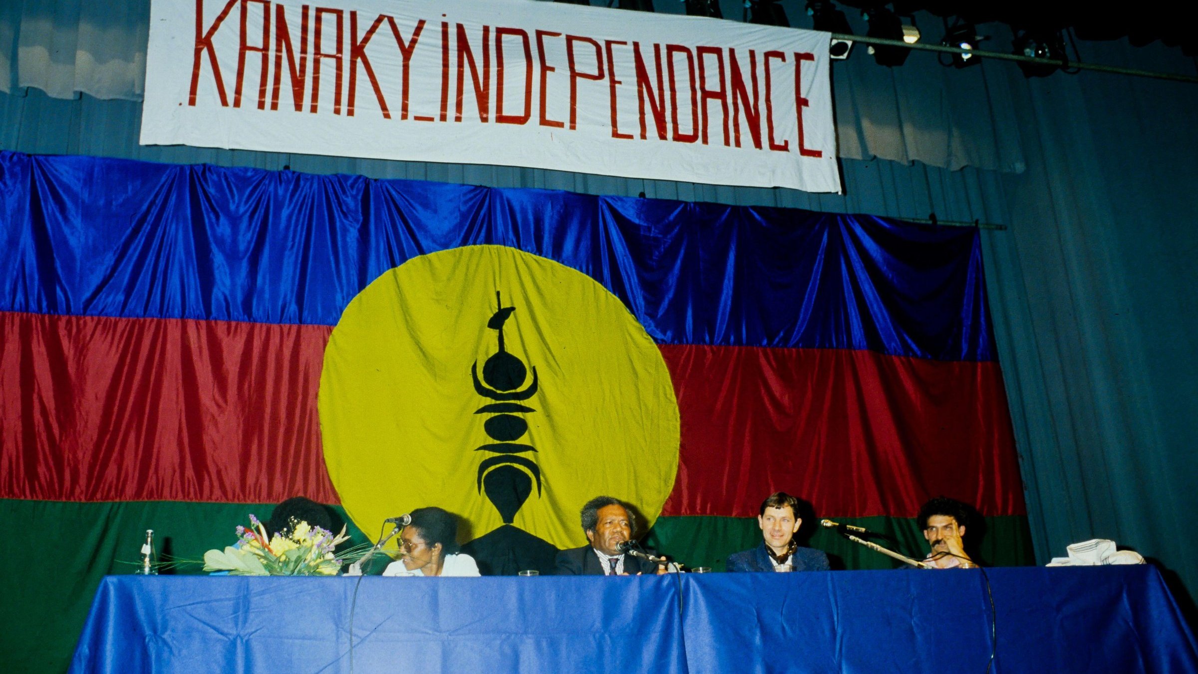 L'image présente une scène de réunion ou de conférence, où des personnes sont assises derrière une table. En arrière-plan, il y a un grand drapeau avec des couleurs vives : un rouge et un bleu, et un cercle jaune au centre. Dans ce cercle, on peut distinguer un symbole noir, précisant ainsi l'identité culturelle. Au-dessus de la scène, une banderole arbore le mot "KANAKY INDEPENDANCE" écrit en lettres rouges, exprimant un message fort lié à l'indépendance. Les participants semblent engagés et sont en discussion, créant une atmosphère de détermination et de solidarité.
