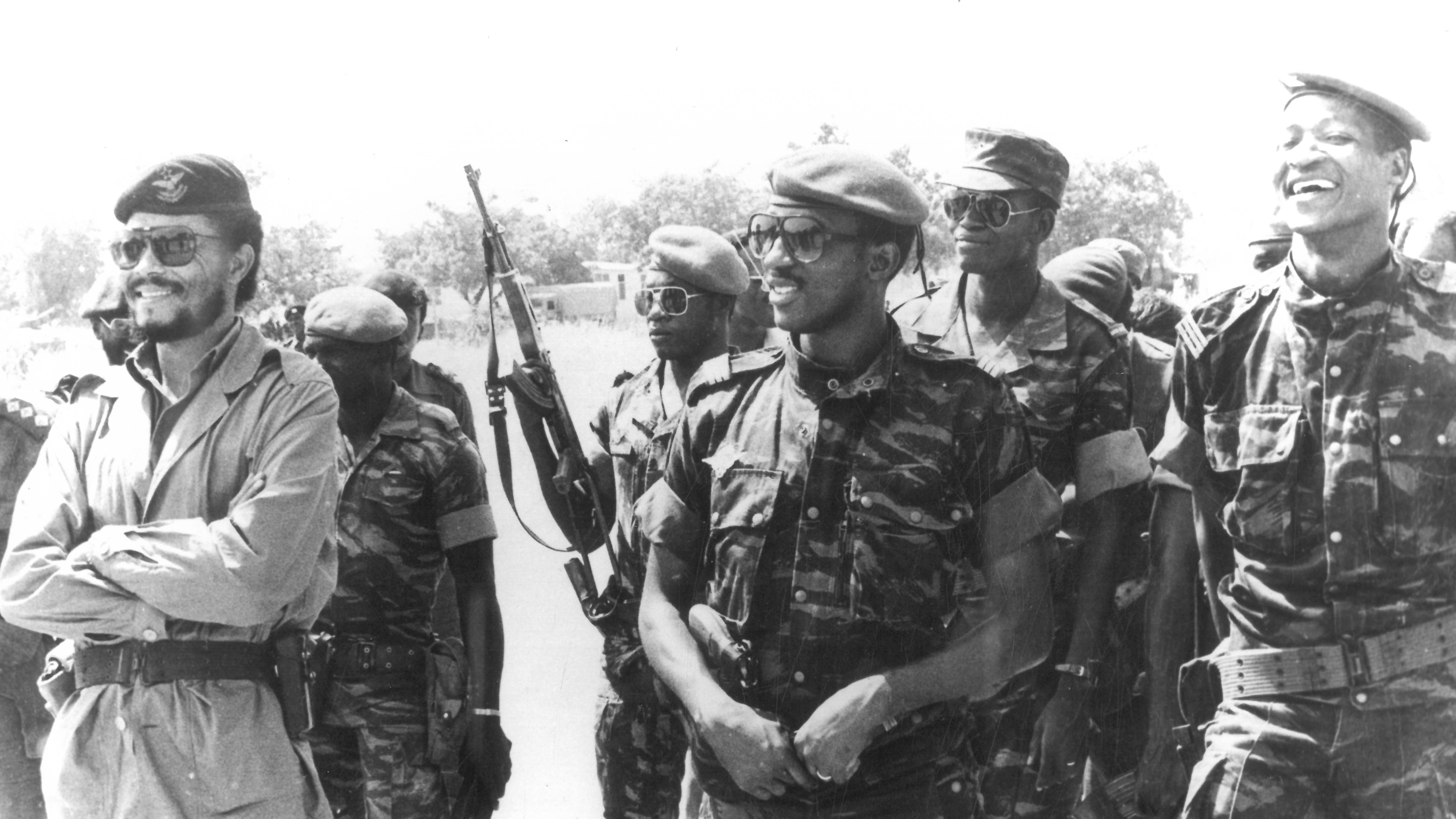 Cette image en noir et blanc montre un groupe de soldats, tous habillés en uniforme militaire. Au centre, un homme souriant porte un beret et un uniforme camouflage. À sa droite, un autre homme, également en uniforme, affiche un large sourire. En arrière-plan, plusieurs soldats tiennent des fusils et portent des lunettes de soleil. L'ambiance semble détendue, avec des expressions de camaraderie parmi les membres du groupe. Le décor en arrière-plan est peu détaillé, mais on peut deviner un environnement extérieur.