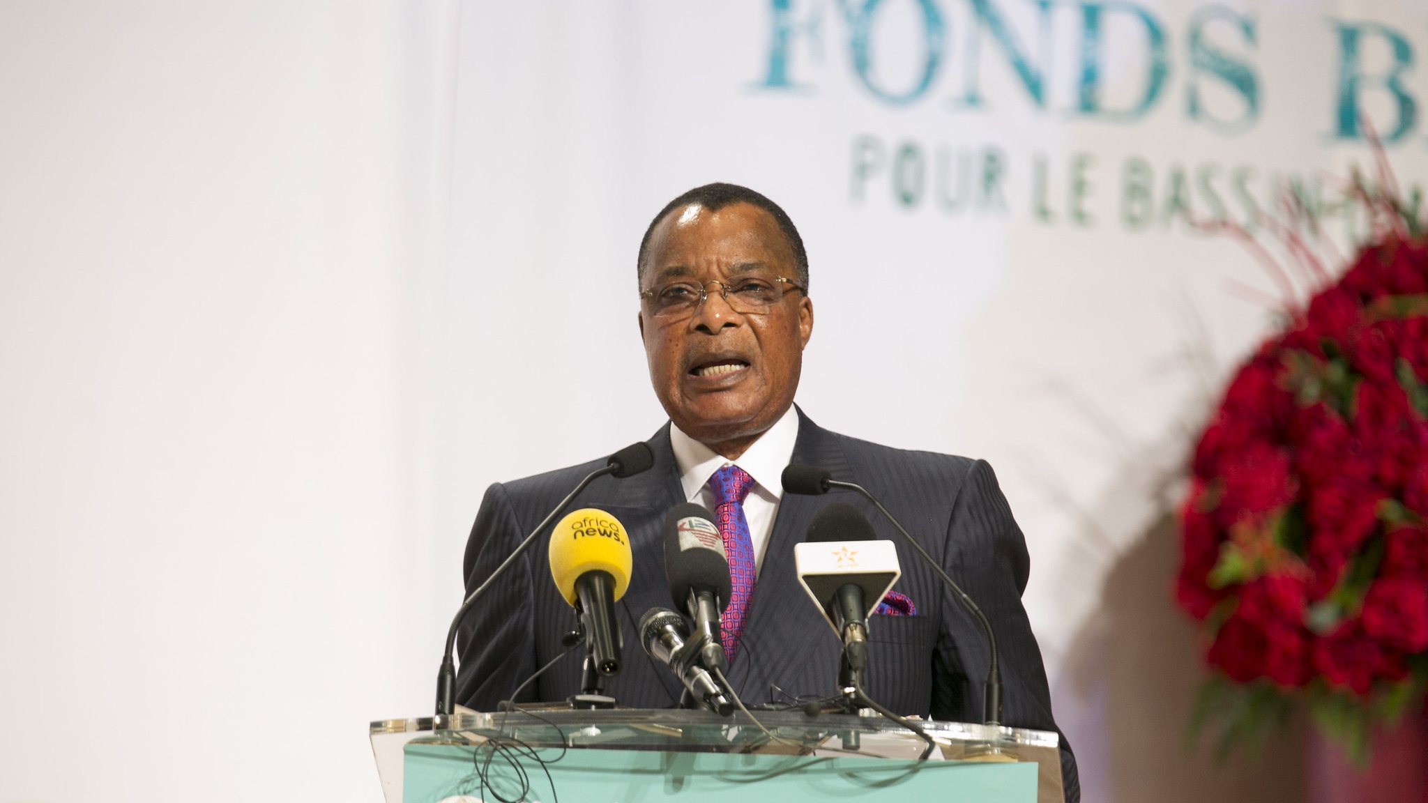 L'image montre un homme, probablement un leader ou un représentant politique, s'exprimant devant un public. Il porte un costume sombre avec une cravate rose et semble être en train de donner un discours. Devant lui, il y a un podium avec plusieurs microphones et un panneau indiquant "Fonds Bleu pour le Bassin du Congo". En arrière-plan, on peut voir des éléments décoratifs qui suggèrent un événement officiel, avec des logos et des affiches liés au thème de la conservation et du développement. L'atmosphère semble sérieuse, indiquant l'importance de l'événement.