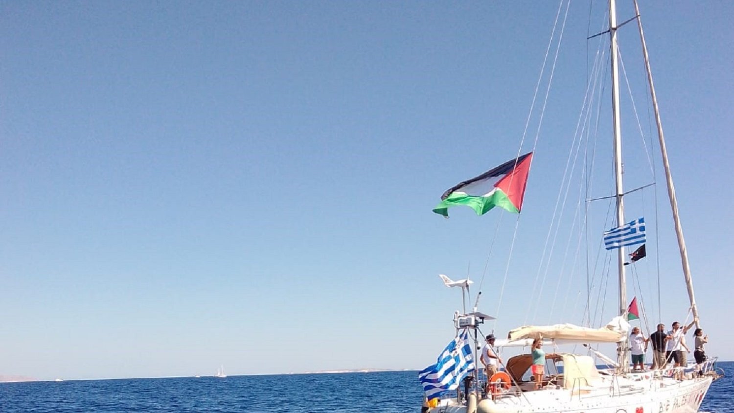 L'image présente un voilier sur une mer turquoise calme, sous un ciel d'un bleu éclatant sans nuages. Le bateau, de couleur blanche, arbore plusieurs drapeaux, dont celui de la Palestine, de la Grèce, et d'autres drapeaux en bandes horizontales typiques. À bord, on aperçoit des personnes qui semblent profiter d'une journée en mer. Les vagues légères créent une ambiance paisible, et l'horizon lointain s'étend, avec d'autres bateaux visibles au loin. L'ensemble évoque une atmosphère de liberté et d'aventure maritime.