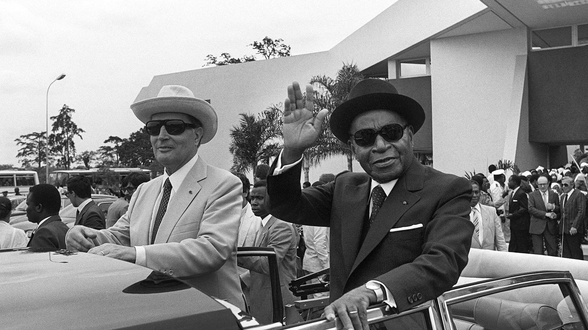 L'image représente deux hommes à l'extérieur d'une voiture décapotable. L'environnement semble être un événement officiel ou une cérémonie, avec des bâtiments modernes en arrière-plan. Le premier homme, à gauche, porte un costume clair, un chapeau de paille et des lunettes de soleil. Il a une expression sérieuse et regardent vers l'avant. Le second homme, à droite, est vêtu d'un costume foncé, d'un chapeau noir et également de lunettes de soleil. Il lève la main en signe de salut, avec un léger sourire. Il y a une foule de personnes en arrière-plan, habillées en costumes formels, ce qui indique une atmosphère festive ou cérémonielle. Des palmiers ou des arbres peuvent être aperçus, ajoutant une touche tropicale à l'environnement.