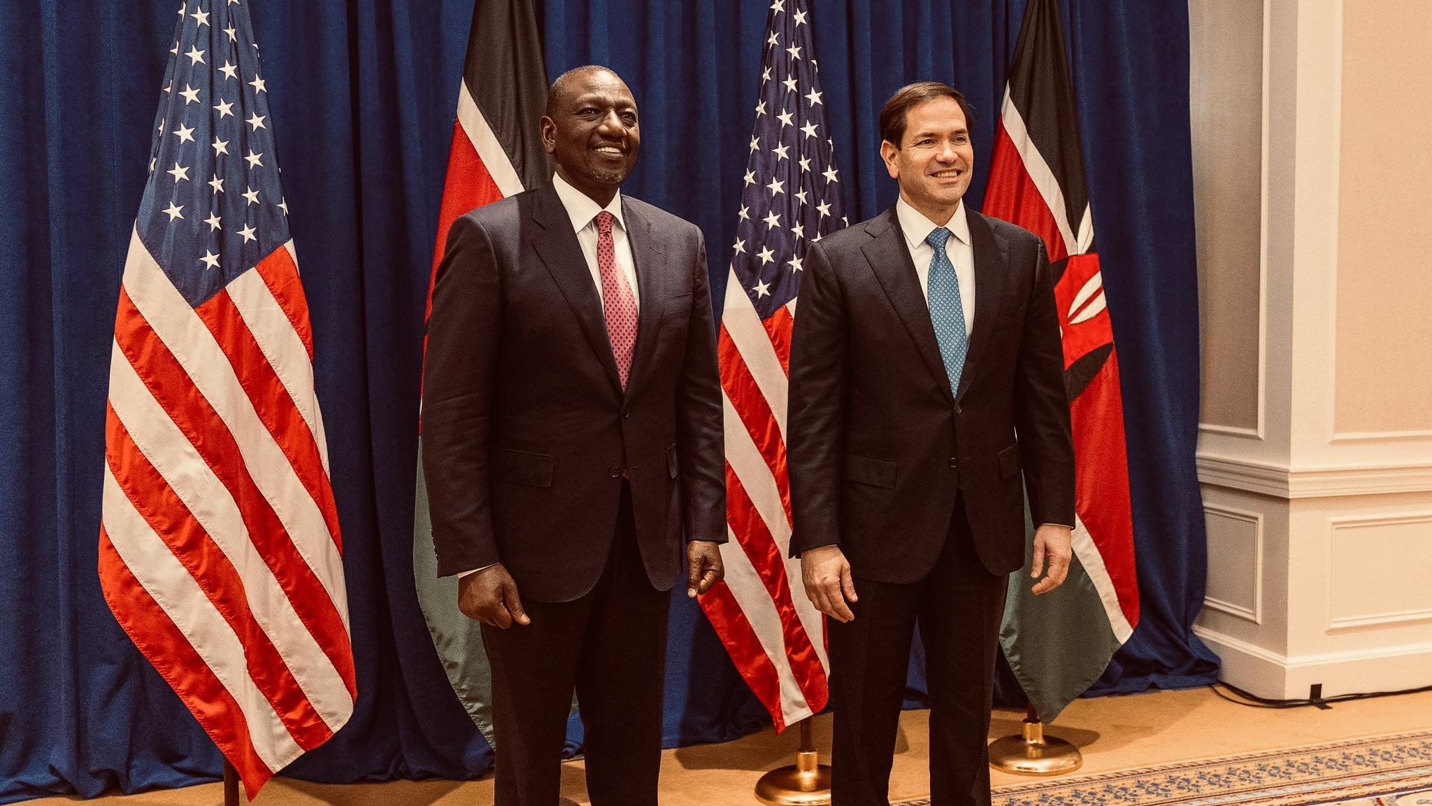 Sur cette image, nous voyons deux hommes debout côte à côte, souriants. À gauche, un homme portant un costume noir et une cravate rouge. Il a la peau foncée et une posture confiante. À droite, un homme avec un costume sombre et une cravate bleu clair. Il a la peau plutôt claire et un sourire engageant. Derrière eux, il y a trois drapeaux : deux des États-Unis, avec des étoiles blanches sur fond bleu et stripes rouges et blancs, et un drapeau du Kenya, qui a des bandes horizontales noires, rouges et vertes, avec un bouclier au centre. Le décor est élégant, avec un fond bleu et un tapis orné à leurs pieds. L'atmosphère semble formelle et amicale.