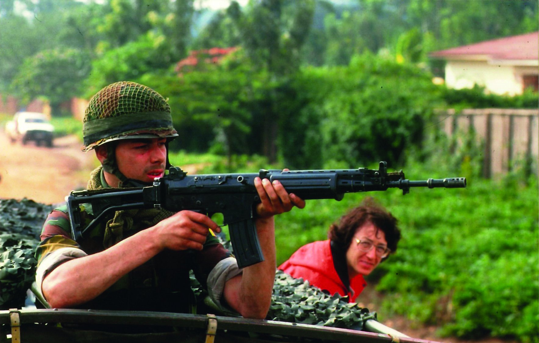 L'image montre un soldat, assis dans un véhicule militaire,tenant un fusil vers l'avant. Il est vêtu d'un uniforme camouflage et porte un casque en treillis. Son visage exprime la concentration et la vigilance, tandis qu'il regarde attentivement son environnement. En arrière-plan, une femme à lunettes observe la scène, semblant préoccupée. La verdure luxuriante entoure le cadre, créant un contraste avec la terre battue sur laquelle se trouve le véhicule. L'atmosphère de l'image est chargée de tension, suggérant un contexte de vigilance et de protection.