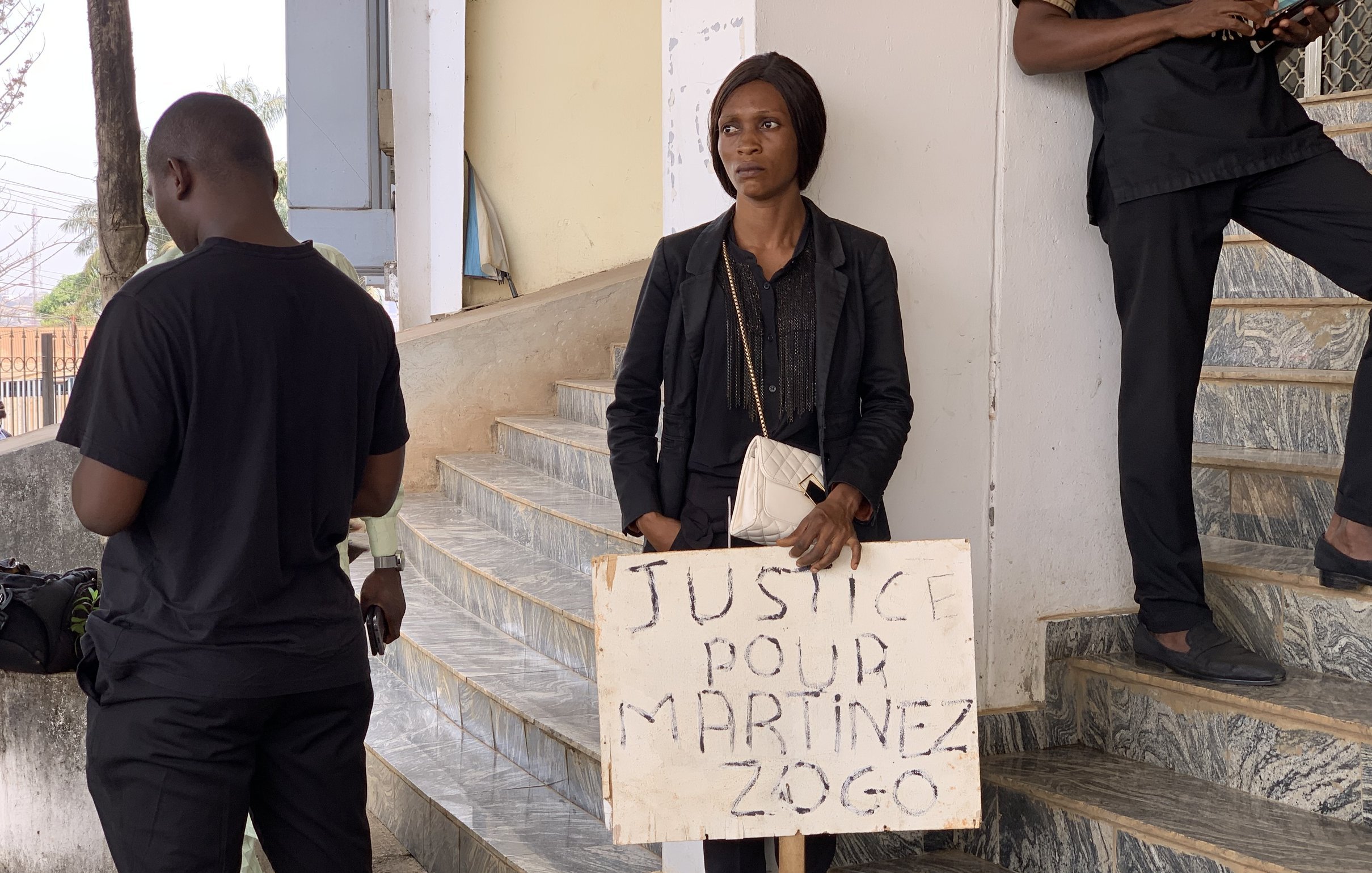 Dans cette image, on voit une femme debout dans un espace public. Elle porte des vêtements noirs et tient un panneau en bois qui porte l'inscription "JUSTICE POUR MARTINEZ ZOGO". La femme a une expression sérieuse et semble déterminée. À proximité, plusieurs personnes en vêtements noirs se tiennent, suggérant un rassemblement ou une manifestation. L'environnement est urbain, avec des escaliers visibles et des bâtiments en arrière-plan, indiquant un cadre où des actions sociales ou des revendications ont lieu.
