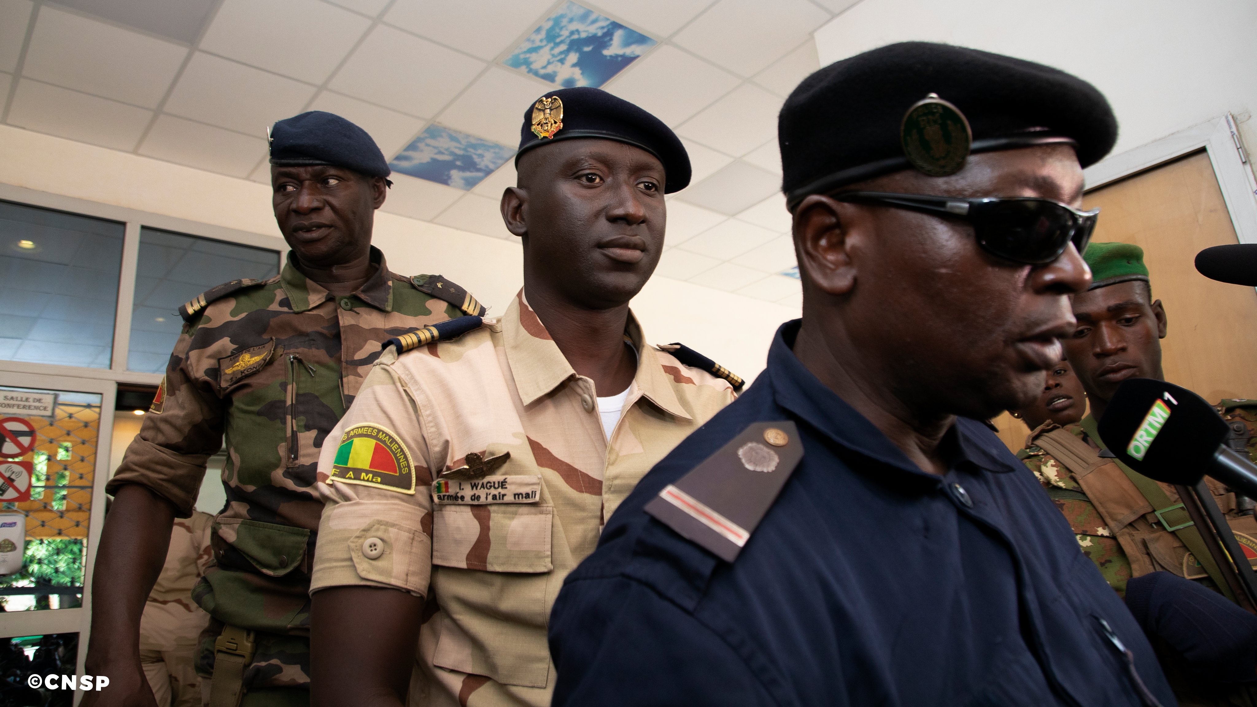 L'image montre un groupe d'hommes en uniforme, probablement des membres des forces de sécurité. Au premier plan, on peut voir un homme en uniforme sombre, portant un chapeau, qui parle à un microphone. Il semble être en train de faire une déclaration ou une interview. À l'arrière-plan, deux autres hommes en uniforme sont présents. L'un d'eux porte un uniforme plus clair avec des insignes, tandis que l'autre a un uniforme de camouflage. L'environnement semble être un bâtiment officiel, avec des murs clairs et un plafond décoré. L'atmosphère peut être perçue comme sérieuse et formelle.