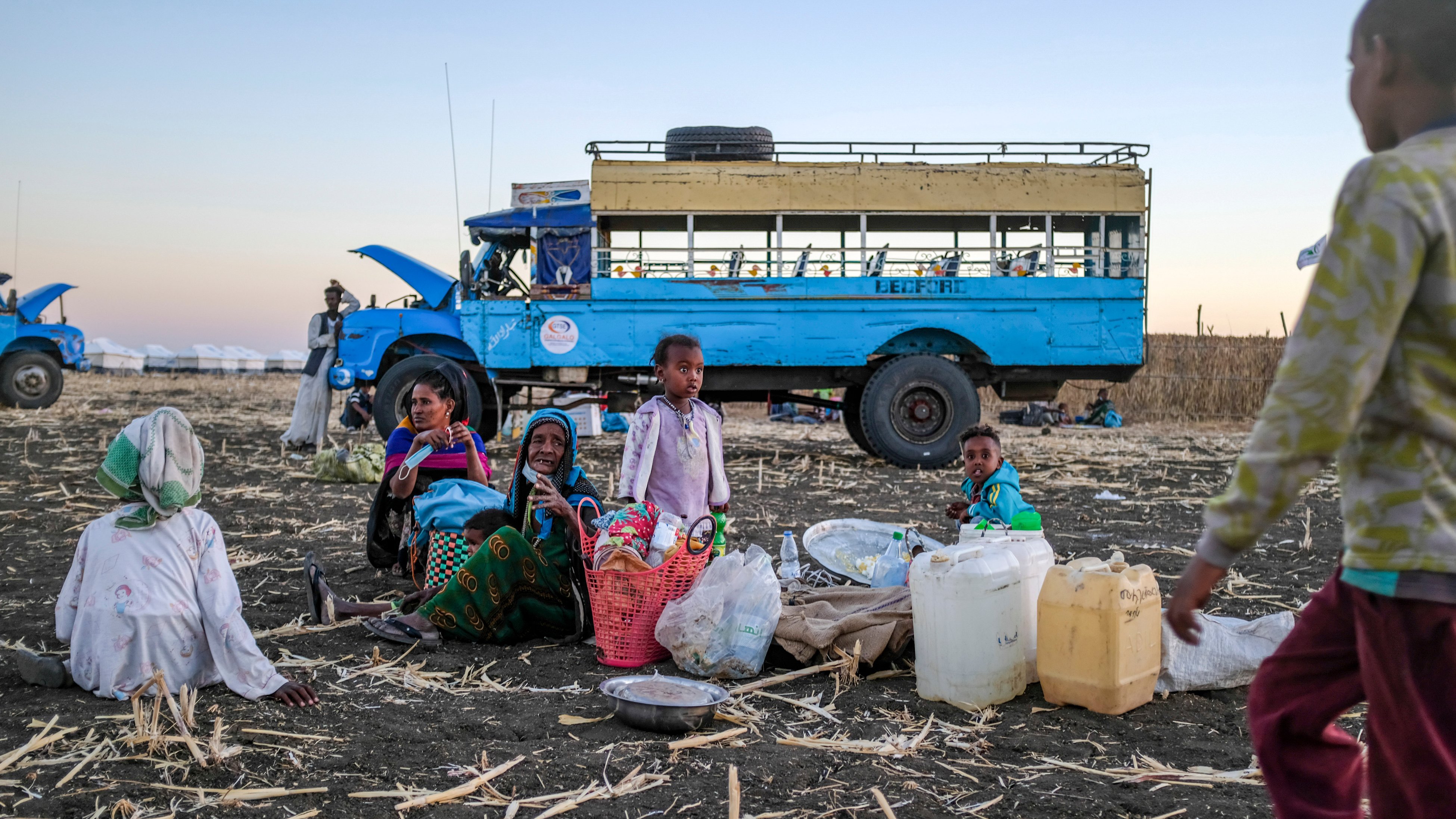 L'image montre un groupe de personnes assises sur un sol de terre battue, entourées de paniers et de contenants. Au centre, des femmes et des enfants semblent converser, tandis qu'un enfant se tient à droite, s'approchant. En arrière-plan, on peut voir un bus bleu immobile, avec un ciel dégradé qui passe des teintes orangées aux bleues, suggérant un crépuscule. Le terrain est jonché de restes de culture, ajoutant un contexte rural à la scène. L'atmosphère évoque une ambiance de rassemblement communautaire dans un environnement de travail.