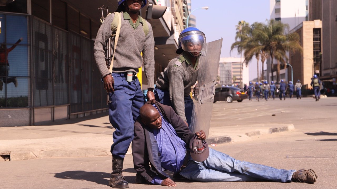 L'image montre une scène de tension dans un environnement urbain. Deux policiers, en uniforme et portant des casques de protection, semblent maîtriser un homme qui est au sol. L'homme est vêtu d'une chemise foncée et d'une veste. Il semble être en difficulté, soutenu par l'un des policiers, tandis que l'autre se tient à ses côtés avec un bouclier. En arrière-plan, on peut apercevoir des bâtiments modernes et quelques palmiers, indiquant que la scène se déroule dans une ville. L'atmosphère générale semble tendue, évoquant un moment de confrontation.