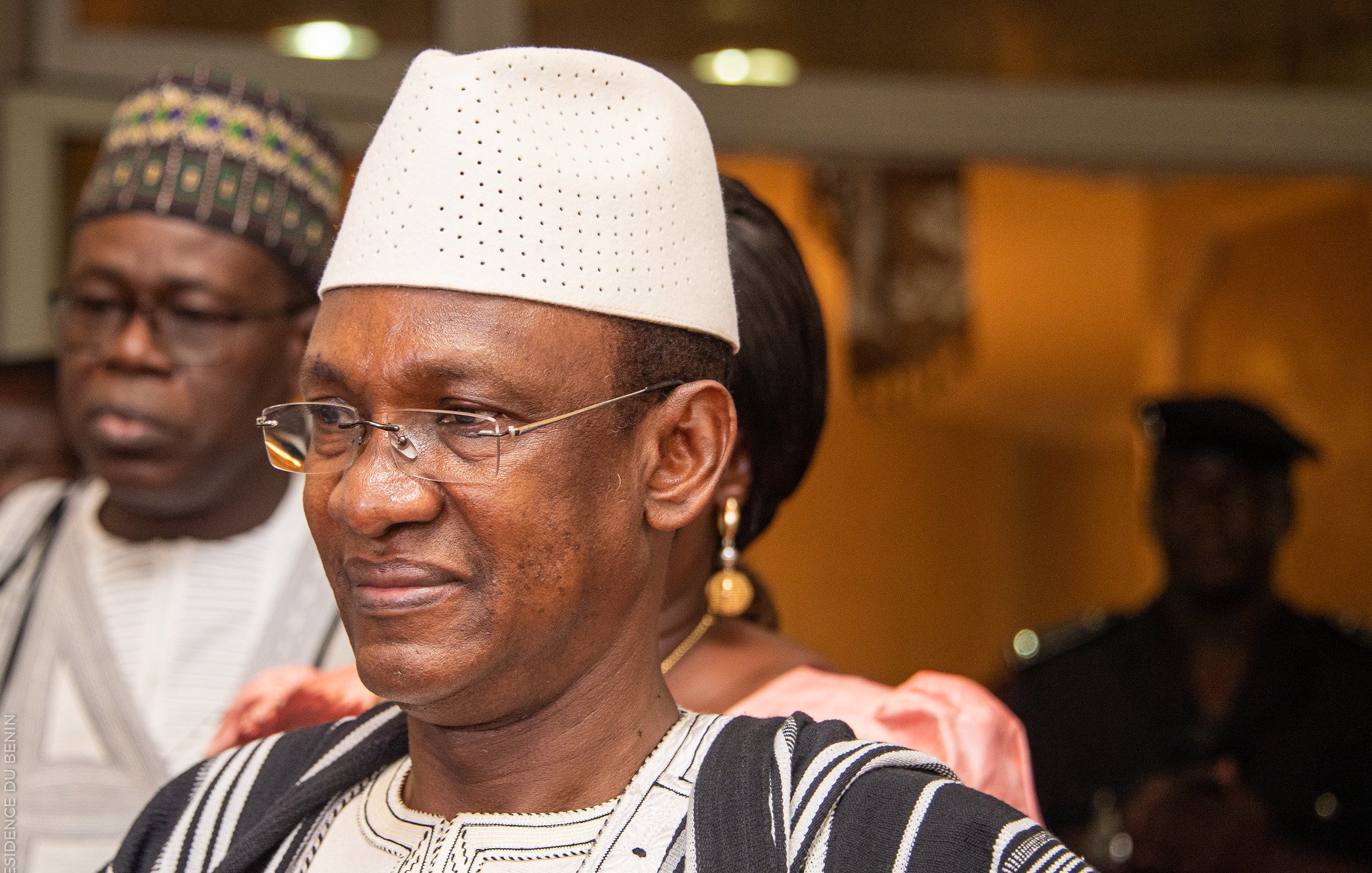 Dans cette image, nous voyons un groupe de personnes dans un cadre formel. L'homme au premier plan porte un chapeau traditionnel blanc et une tenue en tissu bicolore, avec des motifs caractéristiques. Il a un sourire serein sur le visage, illustrant une certaine dignité et bienveillance. À l'arrière-plan, deux autres personnes sont présentes, l'une portant des lunettes et l'autre en tenue traditionnelle également. L'éclairage de la scène suggère une ambiance chaleureuse et accueillante, typique d'un événement social ou culturel.