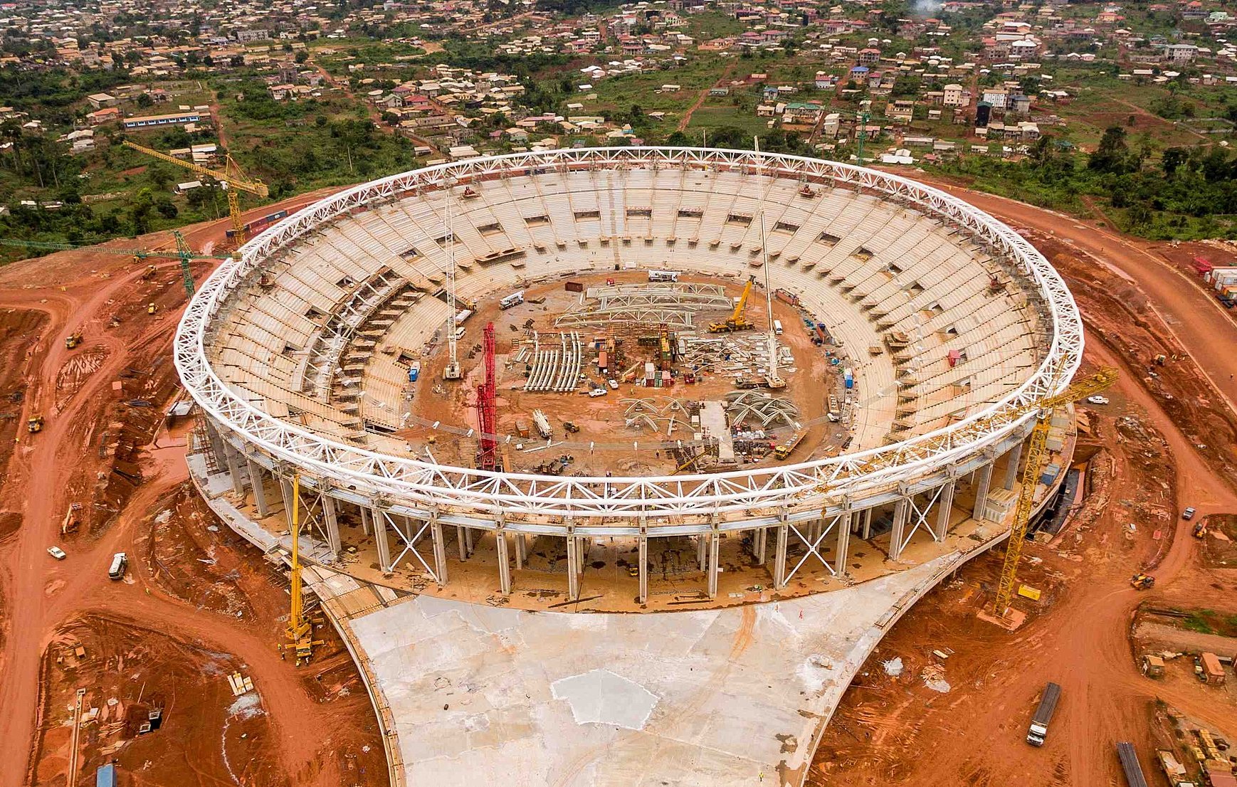 L'image montre un stade en cours de construction, vu d'en haut. Le stade a une forme ovale et un grand espace intérieur qui est encore vide, avec des structures en cours d'installation. Autour du stade, on peut voir un terrain rouge, probablement une terre fouillée pour la construction. À l'extérieur de la structure, il y a des machines de construction et des matériaux éparpillés. À l'arrière-plan, des maisons et des collines sont visibles, ce qui donne une idée de l'environnement urbain et naturel qui entoure le stade. L'ensemble de la scène évoque une ambition de développement et de modernisation.