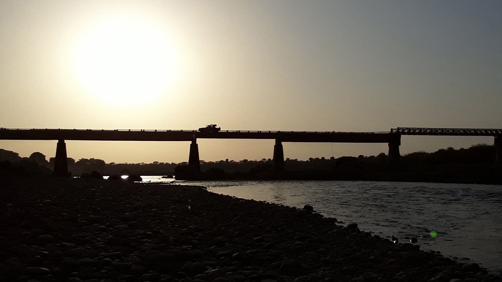 L'image montre un paysage côtier au crépuscule. À l'arrière-plan, on aperçoit un grand pont qui s'étend au-dessus d'une rivière. La lumière du soleil, basse à l'horizon, crée un contre-jour spectaculaire, rendant le pont et les éléments environnants en silhouette. Les contours de la structure sont sombres, se détachant des teintes dorées et oranges du ciel. À proximité de la rive, on peut imaginer des rochers ou des galets, ajoutant une texture au paysage. L'atmosphère est calme et paisible, évoquant un moment de tranquillité près de l'eau.