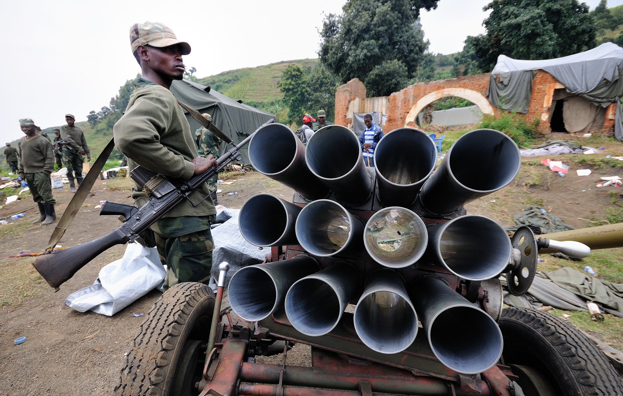 L'image montre un soldat en uniforme, debout à côté d'un véhicule équipé de plusieurs tubes visibles en rangée, qui semblent être des lance-roquettes. Le soldat tient une arme à feu, se tenant avec sérieux. En arrière-plan, on aperçoit d'autres soldats, ainsi qu'un environnement chaotique où des objets sont éparpillés, ce qui donne l'impression d'une zone de conflit ou de préparation militaire. Le tout se déroule dans un paysage verdoyant, avec des collines au loin.