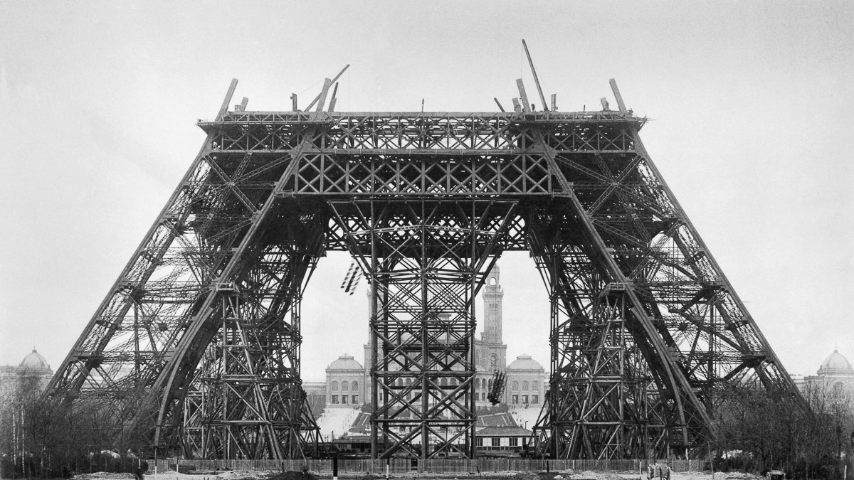 Cette image montre une phase de construction de la célèbre Tour Eiffel à Paris. On peut voir une structure métallique imposante, caractérisée par des poutres en fer entrecroisées. La tour, encore incomplète, présente une base large et stable avec des arcades qui s'élèvent vers le ciel. À l'arrière-plan, on distingue un bâtiment, peut-être une mairie, qui ajoute un contexte urbain à la scène. L'ambiance globale de l'image est monochrome, ce qui renforce l'aspect historique et donne un aperçu des débuts de cette icône de l'architecture. Le sol est encore en chantier, avec des débris et des outils, ce qui rappelle le processus de construction en cours.
