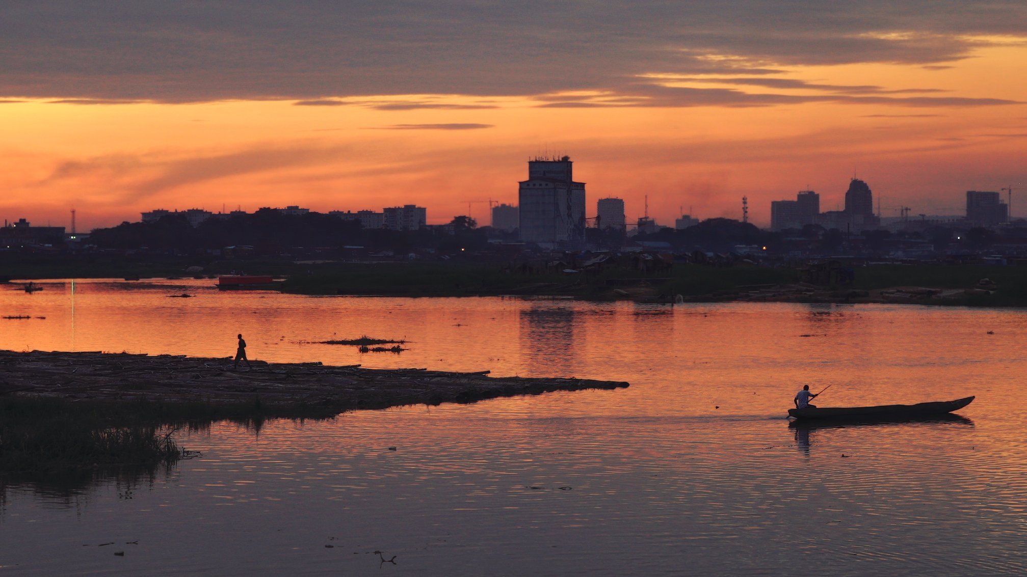 L'image dépeint un paysage paisible au crépuscule. Le ciel est teinté de nuances chaudes d'orange, de rose et de violet, créant une atmosphère calme et sereine. À l'avant, un homme marche au bord de l'eau, tandis qu'un autre navigue tranquillement sur un petit bateau. La surface de la rivière reflète les couleurs du ciel, ajoutant à la beauté de la scène. En arrière-plan, on aperçoit des silhouettes de bâtiments urbains, suggérant la proximité d'une ville. L'ensemble évoque une sensation de tranquillité et de connexion avec la nature, tout en étant en harmonie avec l'activité humaine.