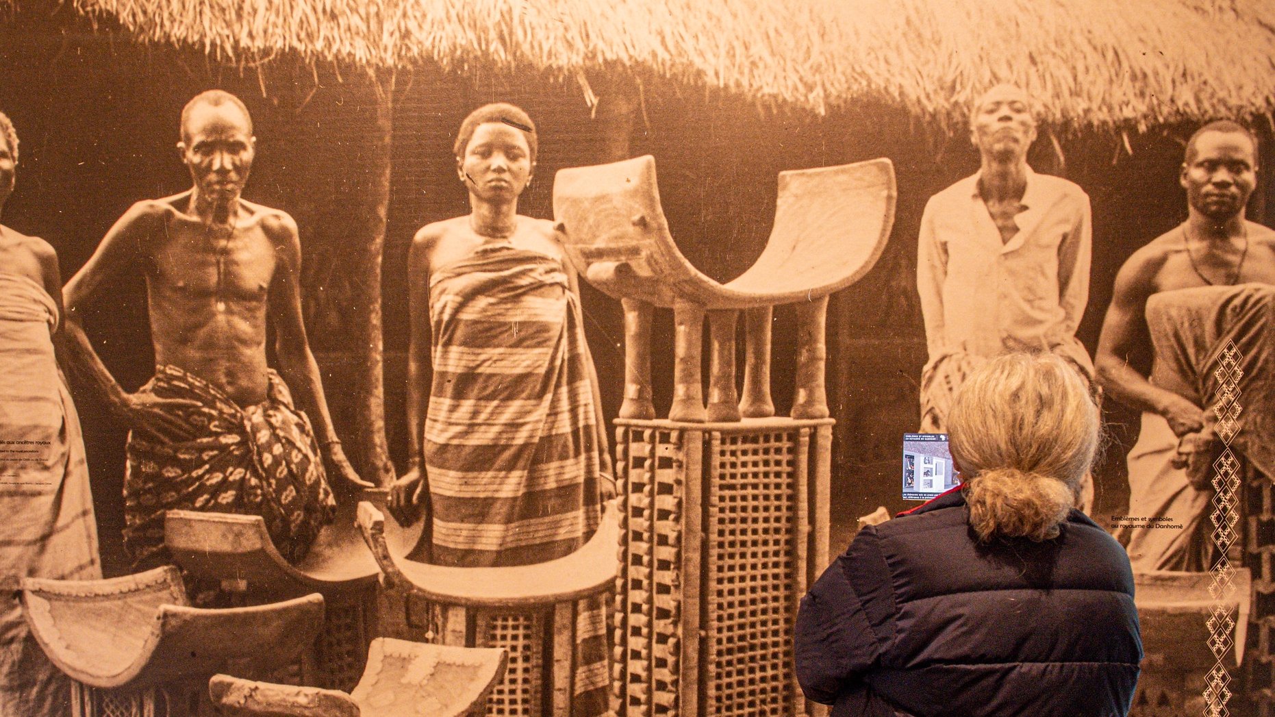 Dans cette image, on aperçoit une scène historique en noir et blanc montrant plusieurs personnes debout, semblant faire partie d'une communauté traditionnelle. Il y a des hommes et des femmes, vêtus de tissus authentiques, avec des poses qui expriment la dignité et la culture. Au centre, on voit des objets artisanaux, comme des chaises ou des tabourets, fabriqués en matériaux naturels, qui portent des motifs décoratifs. À l'arrière-plan, on devine une architecture simple, avec un toit de chaume, créant un cadre authentique. Une femme, de dos, observe cette scène, tenant un appareil dans ses mains, captivée par ce témoignage du passé. L'ambiance générale évoque un sentiment de respect pour les traditions et les histoires des personnes représentées.