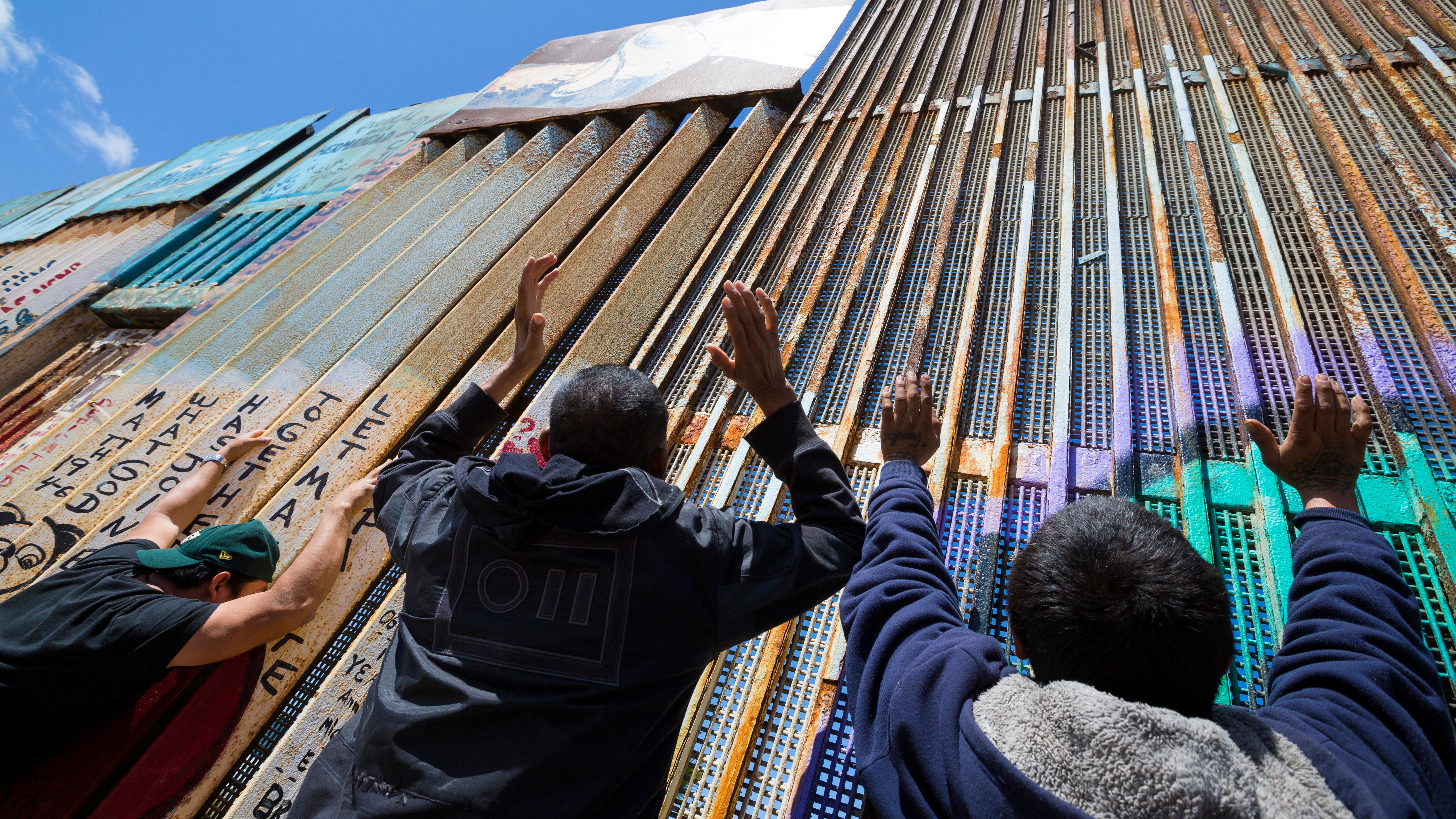 Sur cette image, on peut voir un grand mur en métal qui sépare deux zones. Le mur est composé de panneaux de différentes textures et couleurs, certains recouverts de graffitis. Au premier plan, on aperçoit plusieurs personnes qui tendent les mains vers le mur, comme si elles cherchaient un contact ou un lien. Le ciel est bleu et dégagé en arrière-plan, ce qui crée un contraste avec le métal de la barrière. L'atmosphère semble chargée d'émotion et de désir de connexion.