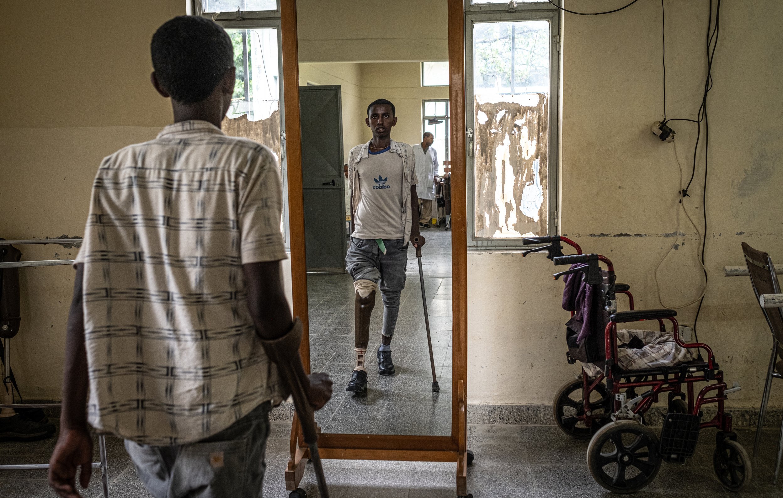 L'image montre une scène dans un lieu qui semble être un centre de réhabilitation ou un hôpital. Au premier plan, un jeune homme se tient à l'aide de béquilles, son regard fixé sur son reflet dans un grand miroir devant lui. Il porte une chemise à rayures et des pantalons clairs. Dans le miroir, on peut voir un autre homme, qui a une prothèse de jambe et se tient debout, semblant plus confiant. Sur le côté, une chaise roulante est disposée, suggérant un environnement d'assistance. Les murs autour d'eux sont simples et montrent des signes d'usure. L'atmosphère évoque des efforts de réhabilitation et de courage.