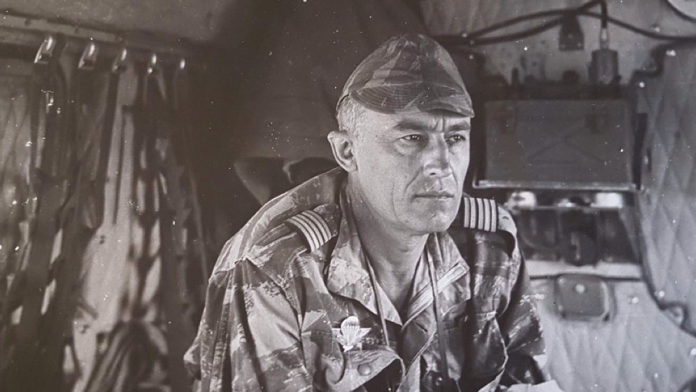 L'image présente un homme en uniforme militaire, assis à l'intérieur d'un véhicule. Il a les cheveux courts et grisonnants, avec une expression sérieuse et réfléchie. Son uniforme, camouflé, est orné d'insignes distinctifs sur les épaules, indiquant son grade. Au fond, on aperçoit des équipements militaires et des matériaux qui suggèrent un environnement de travail opérationnel. La lumière semble douce, et l'atmosphère générale dégage un sentiment de concentration et de détermination.