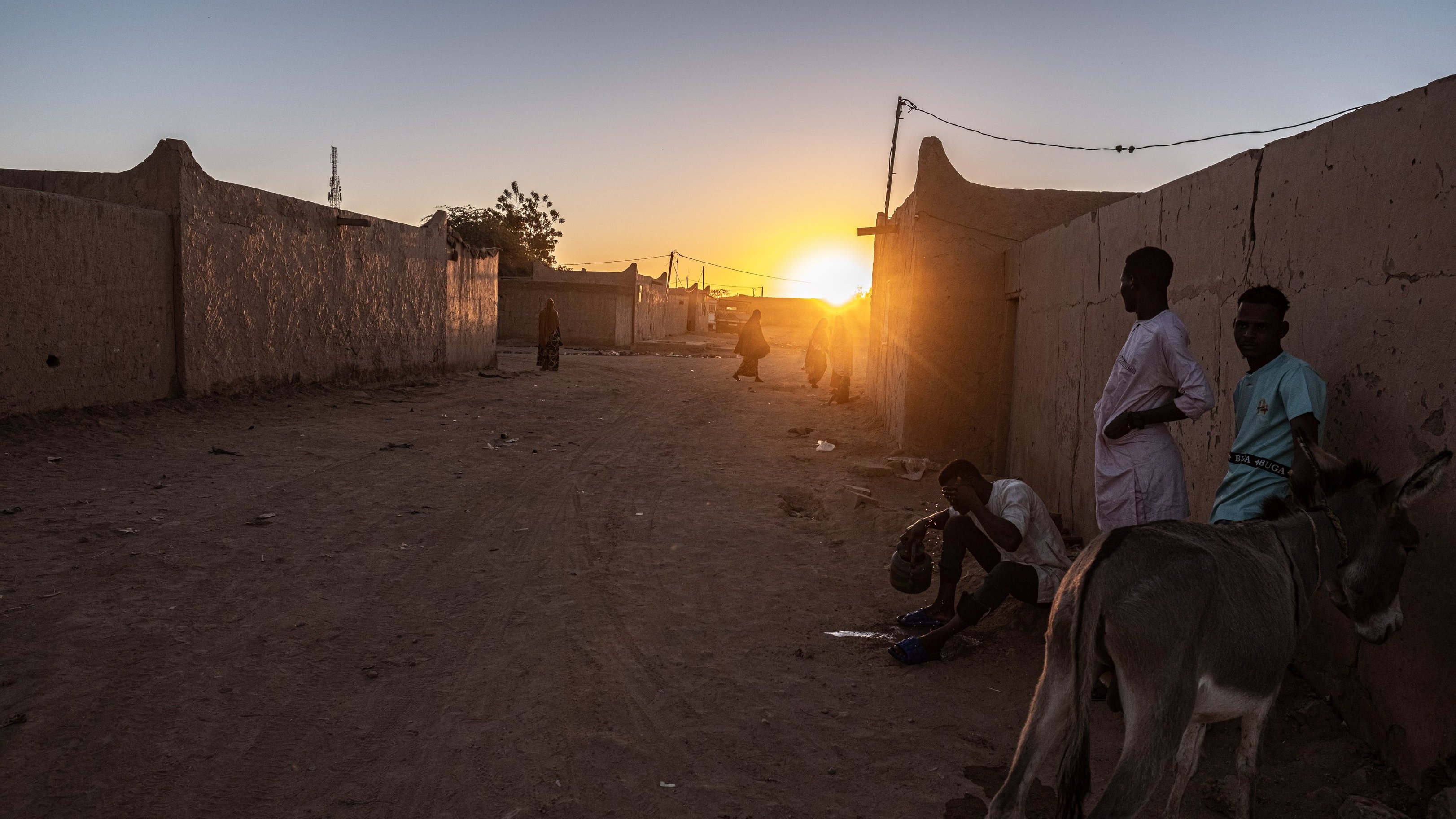 Dans cette image, nous sommes confrontés à un paysage désertique au crépuscule. La lumière du soleil, qui descend à l'horizon, diffuse des teintes chaleureuses d'or et d'orange, illuminant doucement les bâtiments en terre ocre qui bordent la rue. À gauche, un âne se tient paisiblement, ajoutant une touche de vie à la scène. Sur le sol, la poussière et les cailloux témoignent d'une vie quotidienne simple. Des silhouettes d'hommes en vêtements traditionnels se déplacent lentement, discutant ou vaquant à leurs occupations, créant une atmosphère de convivialité et de calme. Les murs des maisons en terre, bien que rudimentaires, dégagent un sentiment de communauté. Dans l'ensemble, cette scène évoque une fin de journée tranquille, où les sons de la nature et les murmures des conversations se mêlent à la douce lumière du crépuscule.