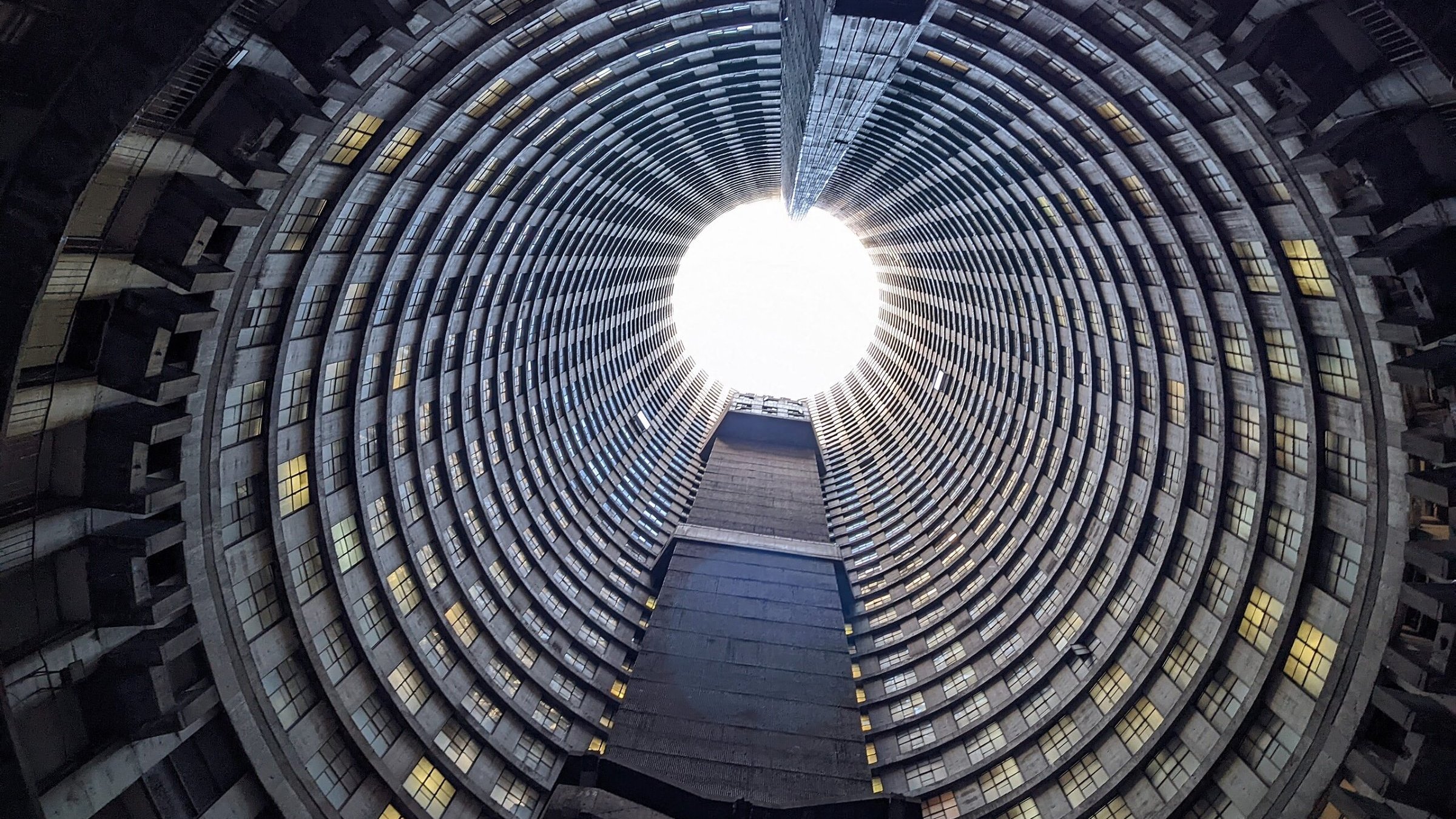 Imaginez vous tenir au centre d'une immense structure circulaire. La vue se dirige vers le haut, où un puits de lumière éclaire l'espace depuis l'ouverture au sommet. Les parois de ce bâtiment sont composées de béton et forment des motifs en spirale, créant une impression de profondeur et de hauteur. Les fenêtres qui bordent les étages, bien que nombreuses, laissent passer une lumière diffuse qui ajoute une sensation d'apaisement. Le sol est plat et peut sembler rugueux, contrastant avec la douceur de la lumière qui découpe le vide en haut. L'atmosphère est à la fois vide et pleine de possibilité, un mélange d'architecture industrielle et de nature, le tout se rejoignant dans ce grand espace ouvert.