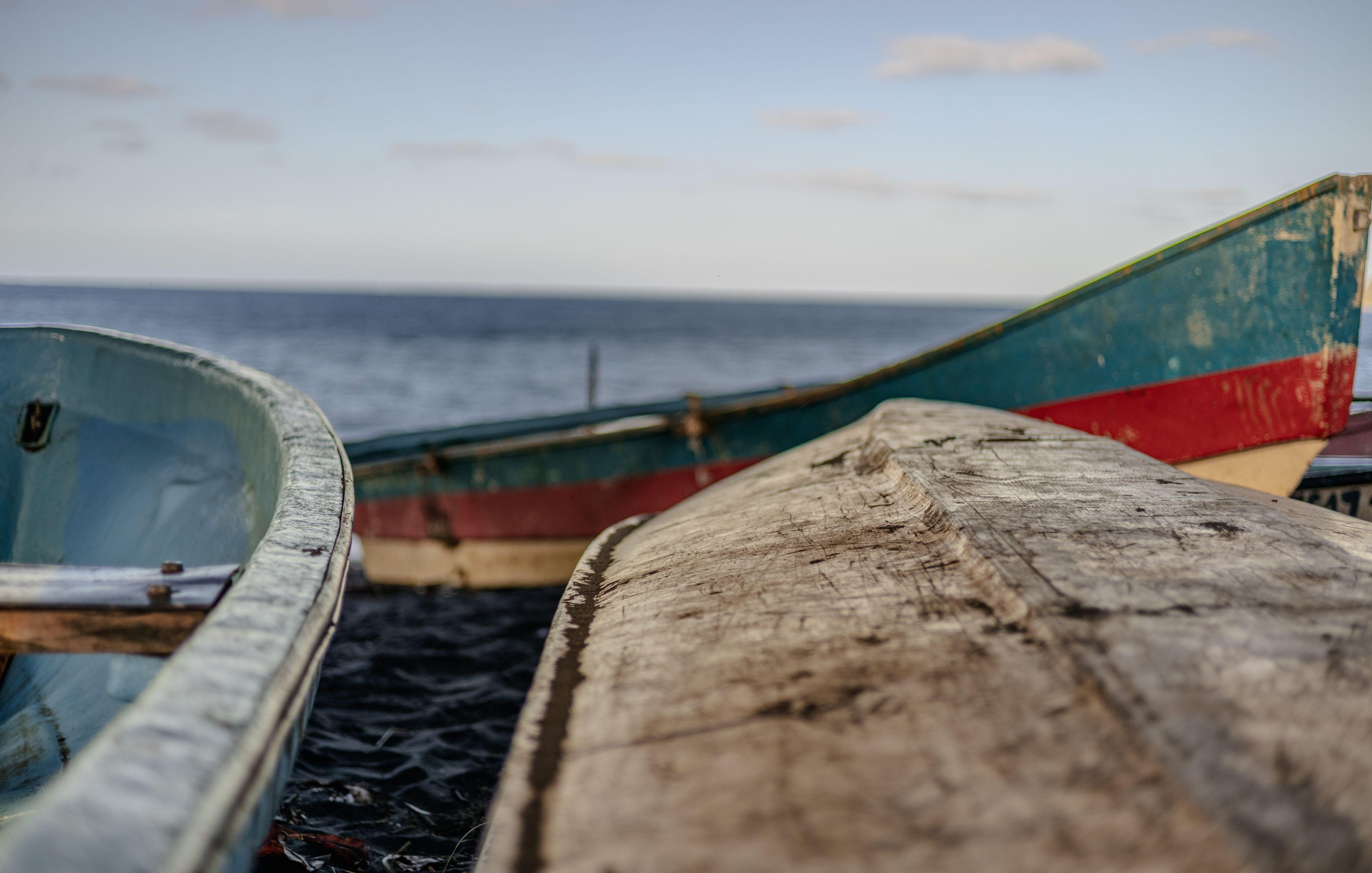 Dans cette image, vous pouvez imaginer un décor paisible au bord de l'eau. Deux bateaux en bois, de designs simples, sont amarrés près d'une plage. Le premier bateau a une structure légèrement arrondie avec une peinture bleu clair sur les côtés, tandis que le second, plus allongé, présente des nuances de bleu et de rouge. Les bateaux sont usés, témoignant de leur utilisation fréquente. En arrière-plan, l'eau s'étend à l'horizon, calme et réfléchissante, et le ciel est dégagé avec quelques nuages légers. L'ambiance est tranquille et maritime, invitant à la contemplation.