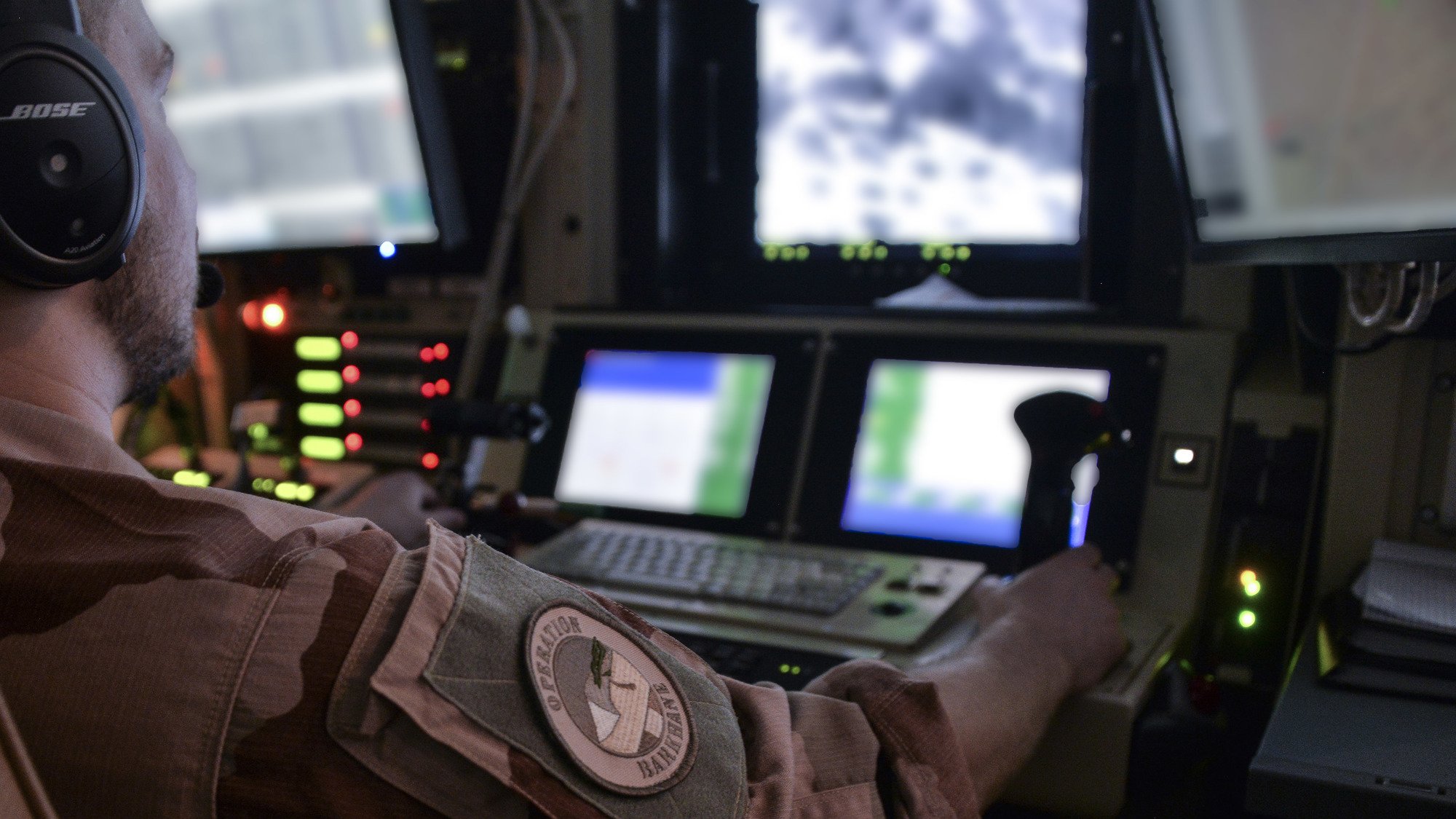 Dans cette image, on voit un homme assis devant un tableau de contrôle, portant un uniforme militaire. Il a un casque sur les oreilles et utilise des manettes et des boutons sur la console. Devant lui, un écran affiche des images en noir et blanc, suggérant qu'il surveille ou contrôle quelque chose à distance. Le décor est sombre, avec des lumières d'indication vertes et rouges autour de lui, ajoutant une atmosphère technologique et militaire à la scène. L'arrière-plan suggère un environnement de travail complexe, probablement dans un avion ou un centre de commandement.