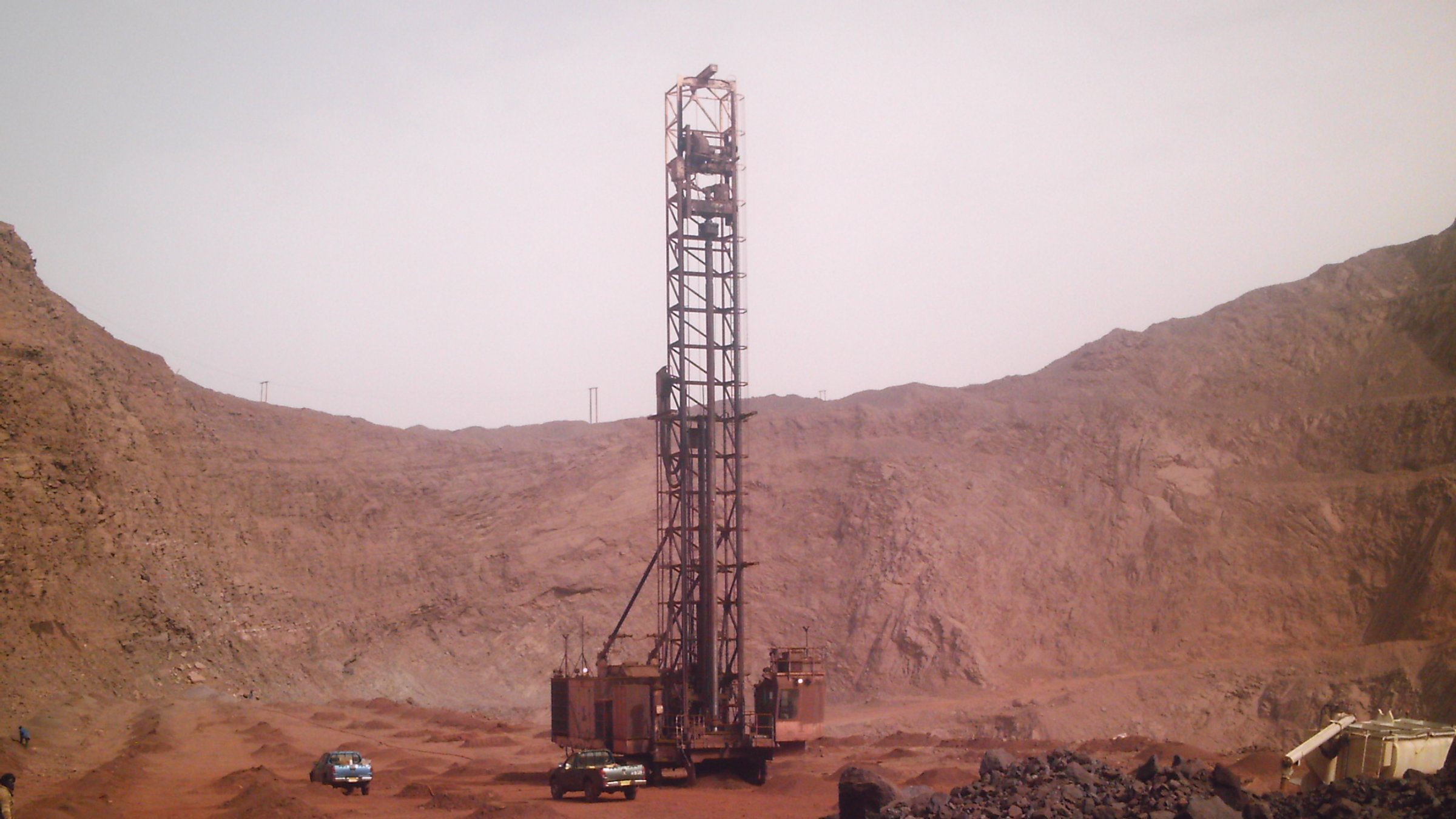 L'image montre un site d'exploitation minière dans un environnement aride et rocheux. Au centre, une grande foreuse se dresse, entourée de montagnes de terre et de roches. La foreuse, de couleur rouille, a une structure métallique imposante qui semble clé dans le processus d'extraction. Autour d'elle, plusieurs véhicules, probablement des camions ou des 4x4, sont garés sur le sol. Le terrain environnant est rocailleux et dépouillé, avec des teintes ocres et brunes, évoquant une atmosphère sévère et industrielle. Le ciel est clair, ce qui accentue l'impression de chaleur et d'aridité de cette zone minière.