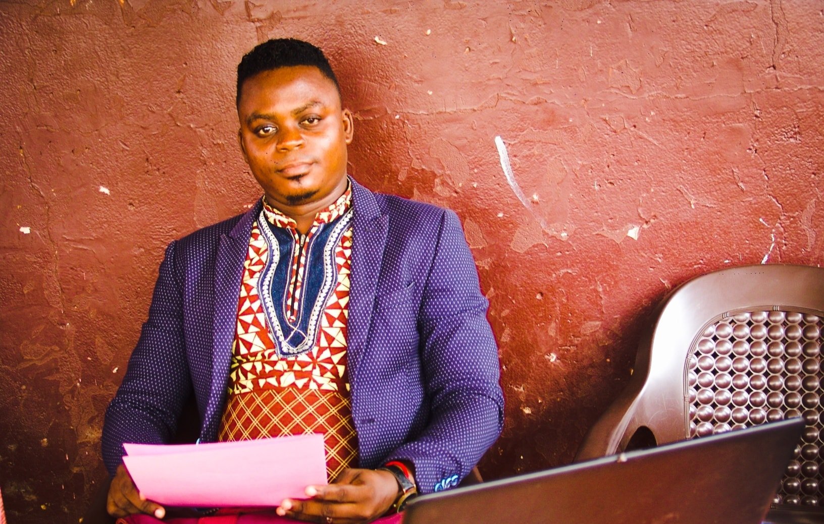 Dans l'image, un homme est assis à une table en bois, près d'un ordinateur portable noir. Il porte une chemise traditionnelle aux motifs colorés, mêlant des nuances de rouge, bleu et blanc, et une veste sombre qui contraste avec sa tenue. Son expression est calme et confiante. Sur la table, on peut voir des feuilles de papier rose ainsi que quelques objets, probablement des lunettes de soleil. Le fond est d'un brun uni, ce qui met en valeur le personnage au premier plan. L'atmosphère semble détendue et conviviale.