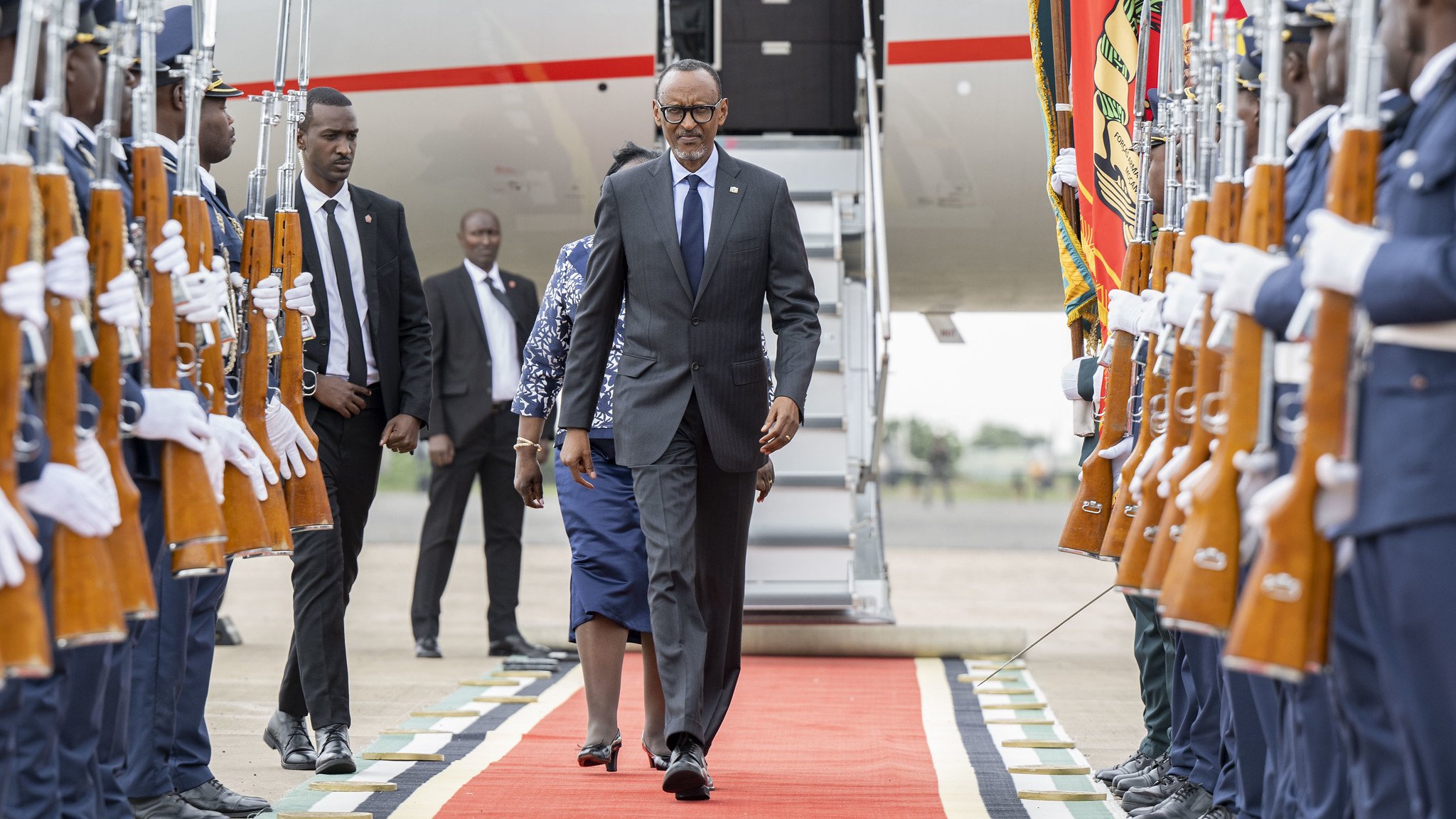 L'image montre un moment officiel sur un tapis rouge, où un homme en costume, probablement un dignitaire, marche avec assurance. Il est entouré de garde d'honneur en tenue formelle, tenant des fusils. À l'arrière-plan, on aperçoit une grande échelle d'avion et d'autres personnes en costume. L'atmosphère dégage un sentiment de cérémonie et de respect, soulignée par la disposition soignée des militaires et le faste du décor.