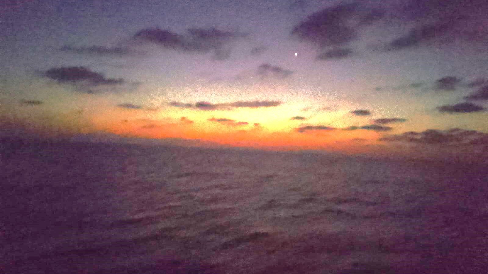 Cette image montre un paysage maritime au crépuscule. Le ciel est teinté de nuances sombres, avec des nuages épars. On remarque un doux dégradé de couleurs allant du bleu foncé au orange, signalant la transition entre le jour et la nuit. L'horizon est légèrement flou, et l'eau semble calme, reflétant les couleurs du ciel. Une étoile brillante est visible dans la partie supérieure de l'image, ajoutant une touche de mystère à cette scène sereine et nocturne. L'atmosphère générale est paisible et contemplative.