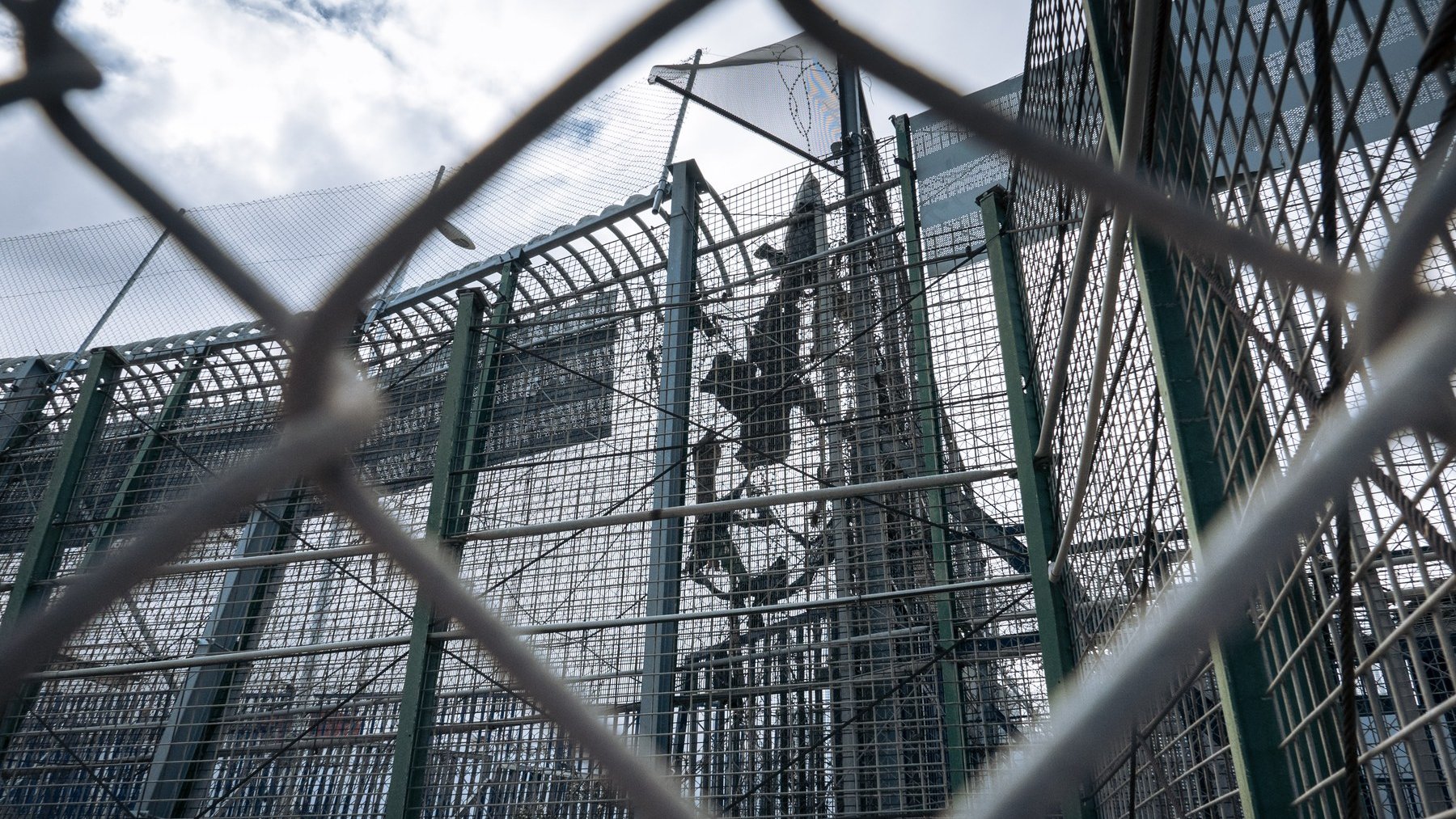 L'image représente une structure de sécurité, probablement une clôture ou un mur de protection. On aperçoit des barrières métalliques en grillage, qui sont transparentes et permettent de voir à travers. À l'intérieur, on distingue une installation, probablement en hauteur, marquée par des éléments solides tels que des poteaux. Le ciel en arrière-plan est nuageux, laissant entendre une lumière diffuse. L'ensemble dégage une impression d'isolement et de confinement, accentuée par la perspective de la clôture.