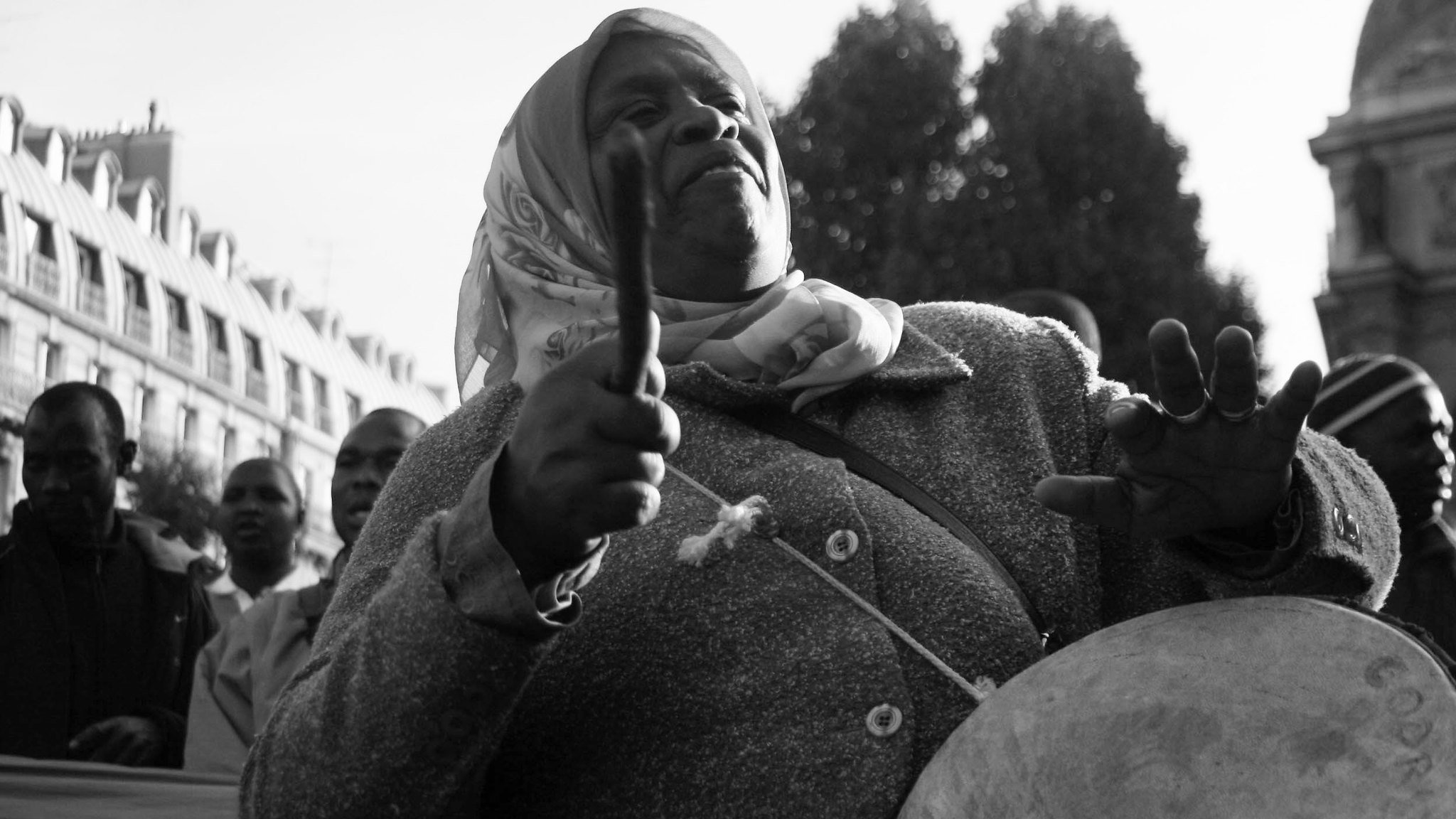 L'image représente une femme engagée dans un moment de mobilisation. Elle porte un manteau gris et un foulard autour de la tête. Ses bras sont levés, tenant des baguettes, et elle semble jouer d'un instrument de percussion, probablement un tambour. Son visage exprime une concentration et une passion profonde, alors qu'elle fait vibrer la musique au sein d'une foule. En arrière-plan, on peut distinguer d'autres personnes, suggérant une manifestation ou un rassemblement collectif. L'atmosphère est chargée d'énergie et de détermination. La photo est en noir et blanc, ce qui ajoute une dimension intemporelle à cette scène de citoyenneté active.