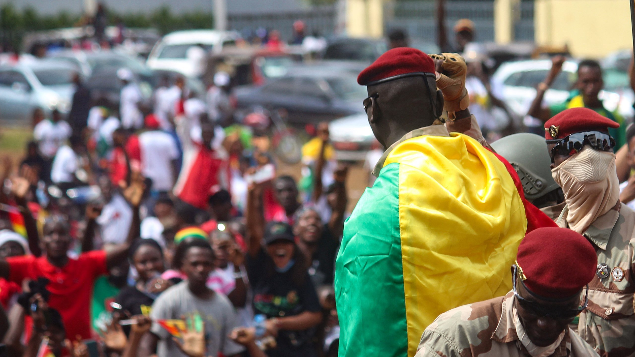 L'image montre une scène dynamique et pleine d'énergie. Au premier plan, un homme en uniforme militaire, portant un béret rouge, se tient sur une plateforme surélevée. Il est enveloppé dans un drapeau aux couleurs vives du Mali, qui est vert, jaune et rouge. Autour de lui, une foule enthousiaste s'agite, brandissant des drapeaux et exprimant leur joie. Les visages des personnes dans la foule montrent des émotions variées allant de l'excitation à la fierté. En arrière-plan, on peut voir des voitures stationnées et d'autres participants se mêlant dans une atmosphère de célébration collective. L'ensemble dégage un fort sentiment d'unité et de patriotisme.