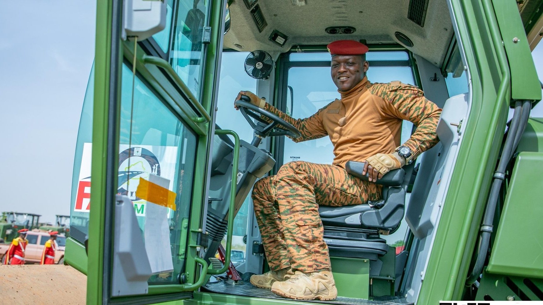 Dans cette image, nous voyons un homme assis à l'intérieur d'un tracteur. Il porte un uniforme couleur sable orné de motifs camouflés et un béret rouge. Son expression est confiante et il sourit, montrant un sentiment de fierté. À l'arrière-plan, on aperçoit d'autres personnes habillées de manière similaire, travaillant dans un environnement agricole. Le ciel est clair et lumineux, suggérant une journée ensoleillée. L'ensemble de la scène dégage une atmosphère de travail collectif et de détermination.