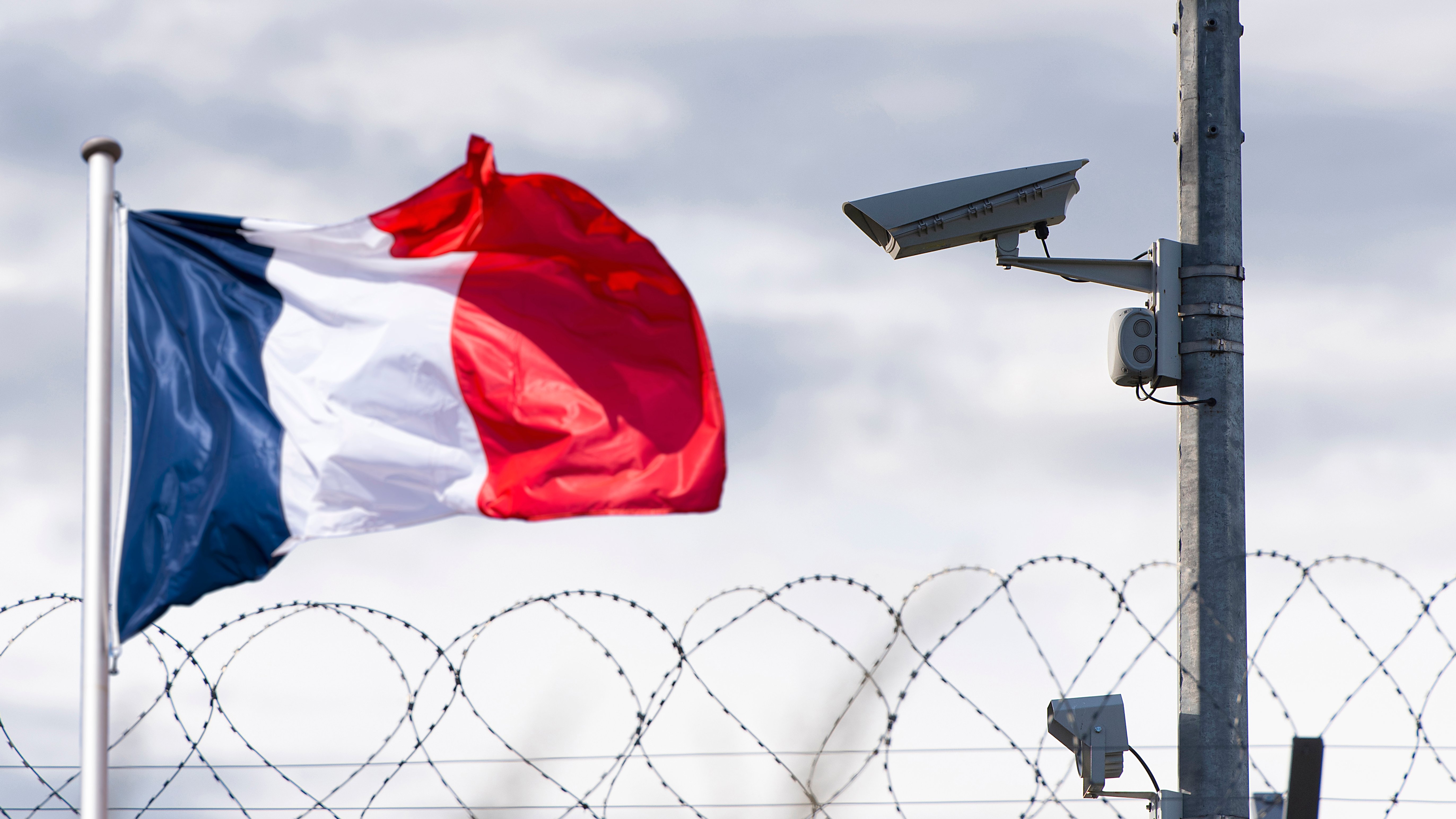 L'image montre un drapeau français vibrant, flottant au vent. À côté, on aperçoit une caméra de surveillance fixée sur un poteau. En arrière-plan, un fil de fer barbelé accentue l'idée de sécurité et de surveillance. Le ciel est nuageux, ajoutant une atmosphère à la scène, qui évoque un environnement protégé.