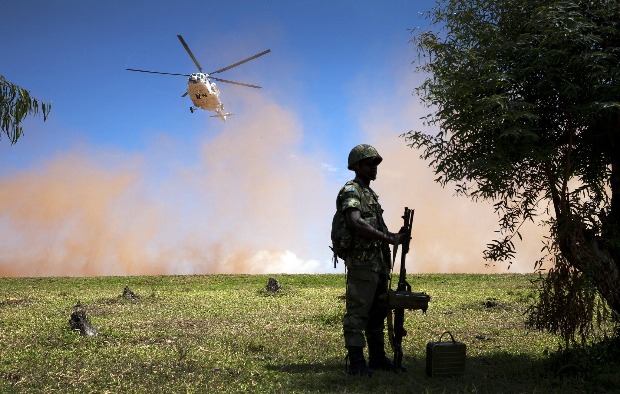 L'image représente un paysage militaire. Au premier plan, on voit un soldat debout, tenant une arme. Il porte un casque et un uniforme camouflage, suggérant qu'il se trouve dans un environnement opérationnel. À l'arrière-plan, un hélicoptère survole la scène, produisant un mouvement et une dynamique. Le ciel est clair, mais il y a également un nuage de poussière ou de fumée, ajoutant une atmosphère dramatique à la scène. Le terrain est verdoyant, mais des éléments comme des troncs d'arbres ou des obstacles peuvent être perçus, contribuant à l'ambiance militaire et à l'action en cours.