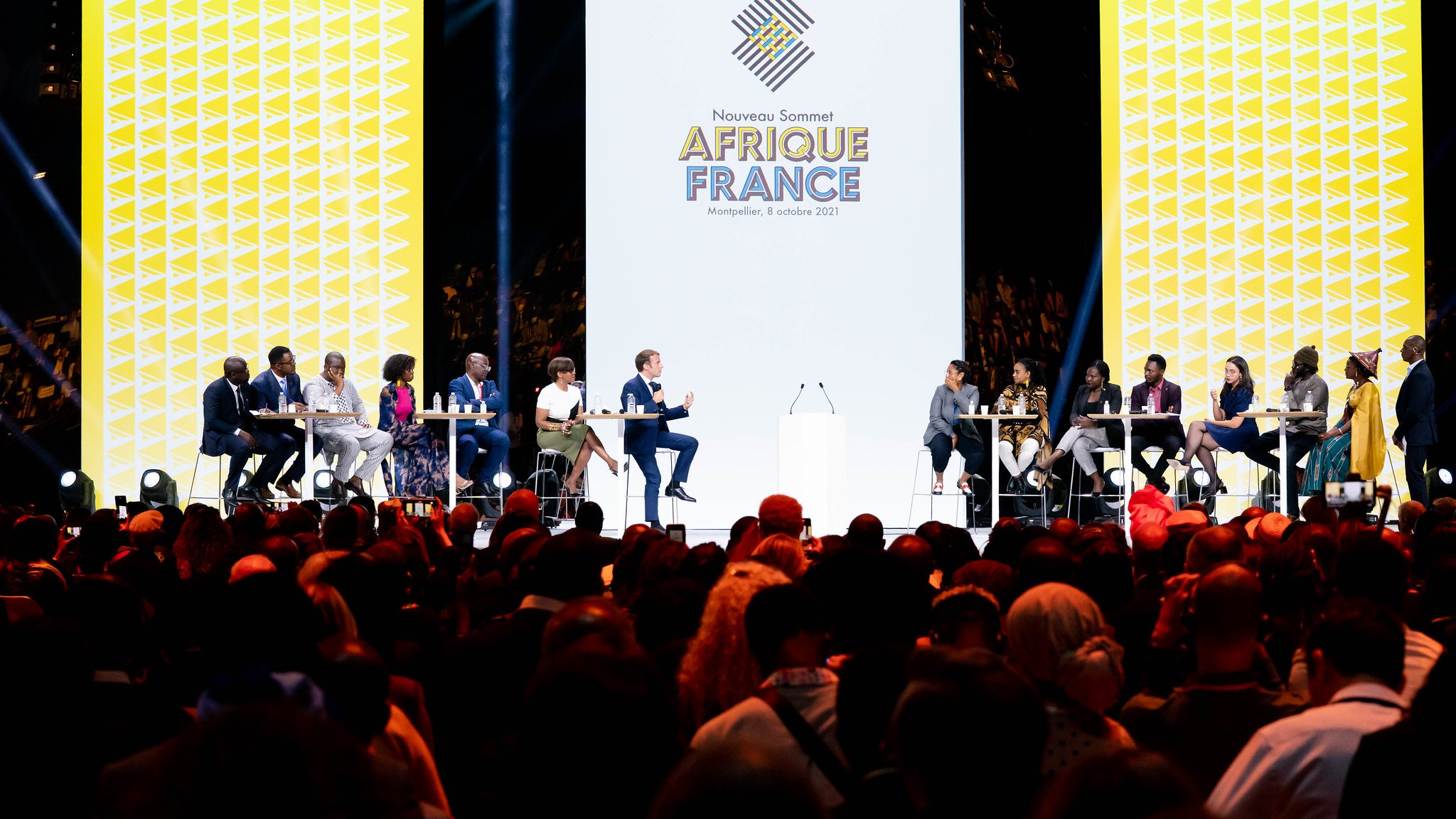 L'image montre une scène d'événement avec plusieurs intervenants assistant à un sommet intitulé "Afrique France". Au centre, un grand écran affiche le logo de l'événement avec une conception graphique moderne. Les intervenants, assis à des tables en cercle, semblent engagés dans une discussion. L'audience est nombreuse et variée, occupant une grande partie de l'espace devant la scène, créant une atmosphère dynamique et interactive. L'éclairage est focalisé sur la scène, mettant en valeur les participants.