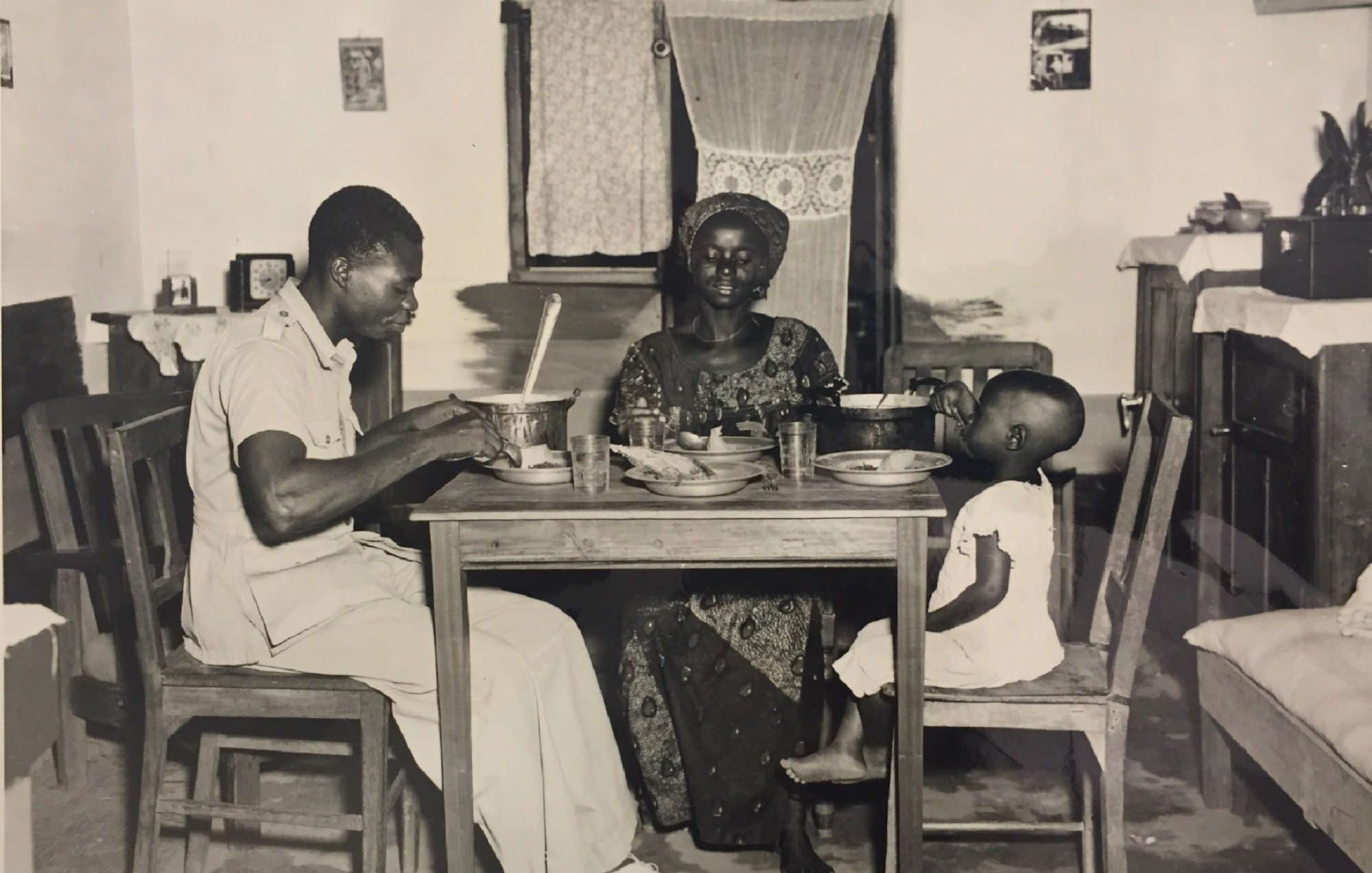 L'image en noir et blanc montre une scène de repas familial dans une pièce simple, probablement une cuisine. À une table en bois, un homme en uniforme, assis sur une chaise, semble être en train de manger avec une grande cuillère. En face de lui, une femme, vêtue d'une robe traditionnelle et d'un chapeau, regarde avec un sourire bienveillant. À côté d'elle, un petit enfant est assis sur une autre chaise, portant une petite robe blanche, observant la scène. Les murs de la pièce sont décorés sobrement, et il y a des verres sur la table, ajoutant une touche de quotidien à ce moment plein de convivialité.