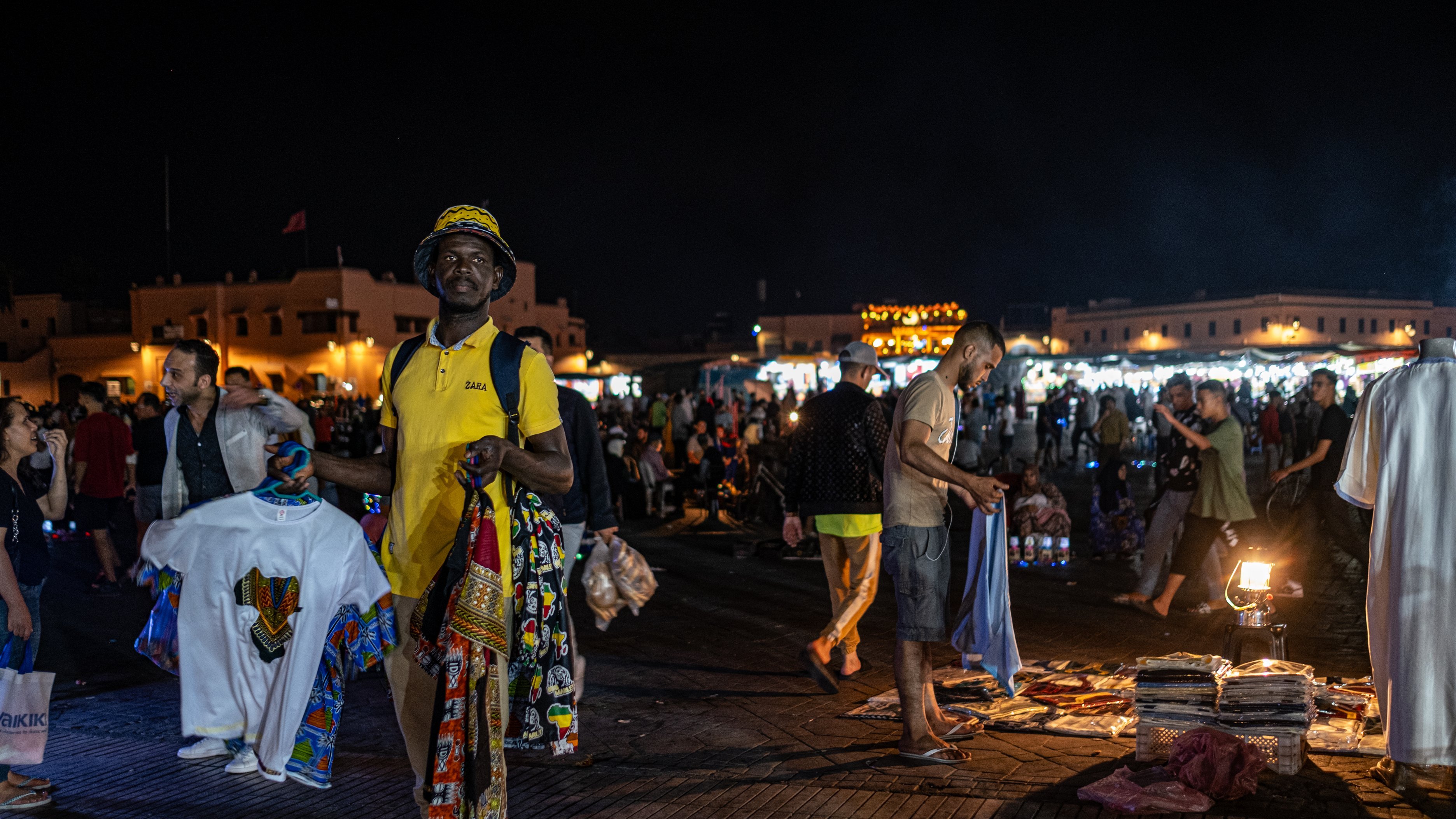 Dans cette image prise la nuit dans un marché animé, on voit un homme au premier plan portant un t-shirt jaune et un chapeau. Il tient plusieurs vêtements, avec des motifs colorés, dans chaque main. L'arrière-plan est rempli de personnes se déplaçant dans le marché, créant une ambiance vivante et dynamique. On peut également apercevoir des lumières brillantes provenant de stands qui éclairent la scène, ajoutant à l'animation nocturne du lieu. La texture du sol semble être en bois ou en pavé, créant un environnement chaleureux. L'air est rempli des sons de conversations et de l'agitation du marché.