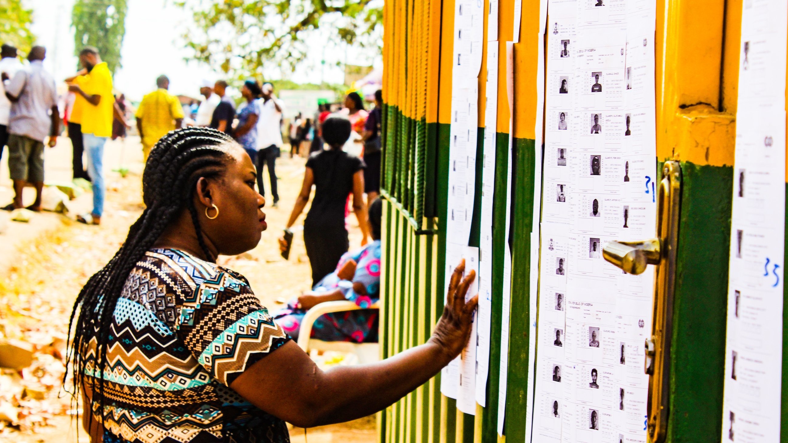 Dans cette image, on voit une scène animée en extérieur. Une femme, avec des tresses et portant un haut aux motifs colorés, se tient près d'un mur recouvert de plusieurs feuilles de papier. Elle passe sa main sur l'une de ces feuilles, qui semble contenir des informations, probablement des listes ou des résultats. En arrière-plan, de nombreuses autres personnes se déplacent, créant une atmosphère de rassemblement. Les couleurs vives du mur et la lumière du jour ajoutent une ambiance chaleureuse à la scène. Au sol, on peut apercevoir des feuilles mortes, ce qui suggère une saison automnale.
