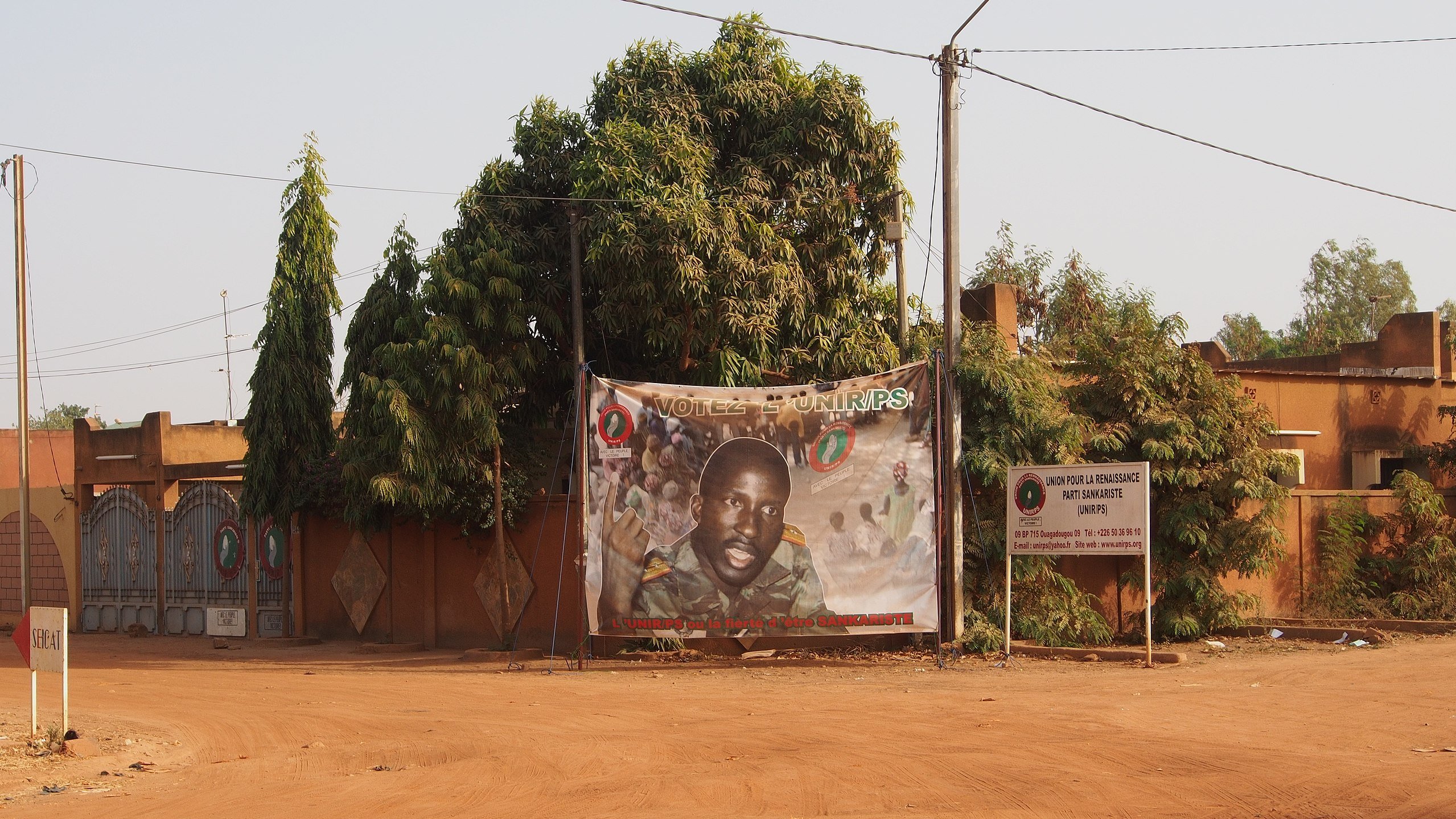 L'image présente une rue déserte d'un quartier, où le sol est recouvert de terre ocre. À gauche, un grand arbre luxuriant, avec des feuilles vertes, apporte une touche de nature. Au centre, une affiche enroulée sur un grillage présente le portrait d'un homme, probablement un candidat politique, souriant et regardant vers l'objectif. L'affiche est ornée de divers motifs colorés et de slogans. À droite, on peut distinguer un panneau d'information sur les élections. Les murs en arrière-plan sont peints dans des tons chauds, contribuant à l'atmosphère locale et authentique de l'endroit.