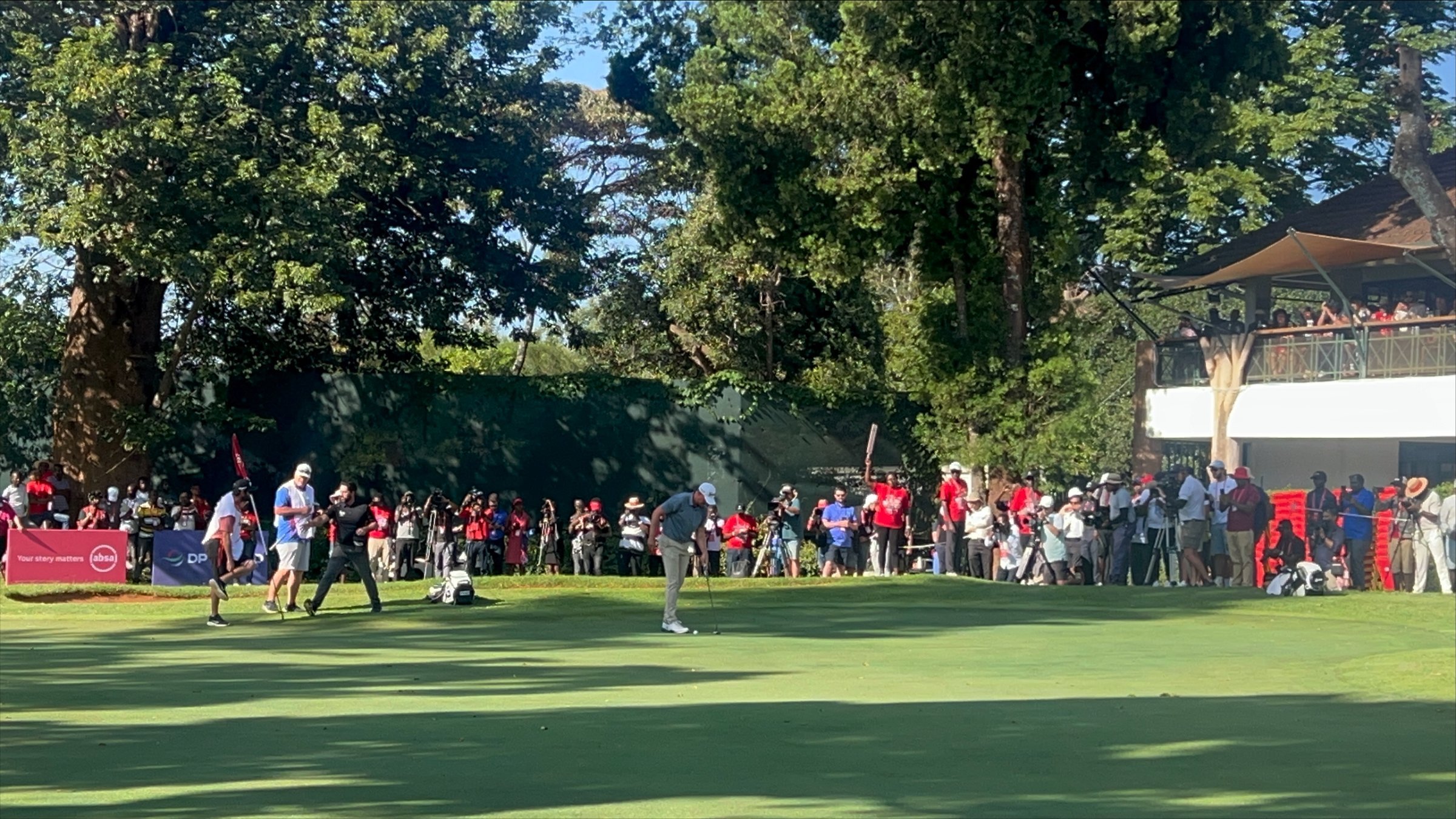 Imaginez un vaste terrain de golf, baigné par la lumière du soleil. Au premier plan, deux joueurs se concentrent sur le putting green, faisant preuve de délicatesse et de précision. L'un d'eux est en position de putt, tandis que l'autre observe attentivement. À l'arrière-plan, des spectateurs en tenue décontractée, vêtus de couleurs variées, attendent avec excitation, certains filmant ou prenant des photos. Autour du green, de grands arbres fournissent de l'ombre, ajoutant à la beauté de l'environnement. À droite, on aperçoit un bâtiment avec des balcons, où d'autres personnes sont rassemblées pour profiter du spectacle. L'atmosphère est vivante, remplie de murmures, de rires et de l'anticipation d'un moment décisif dans le jeu.