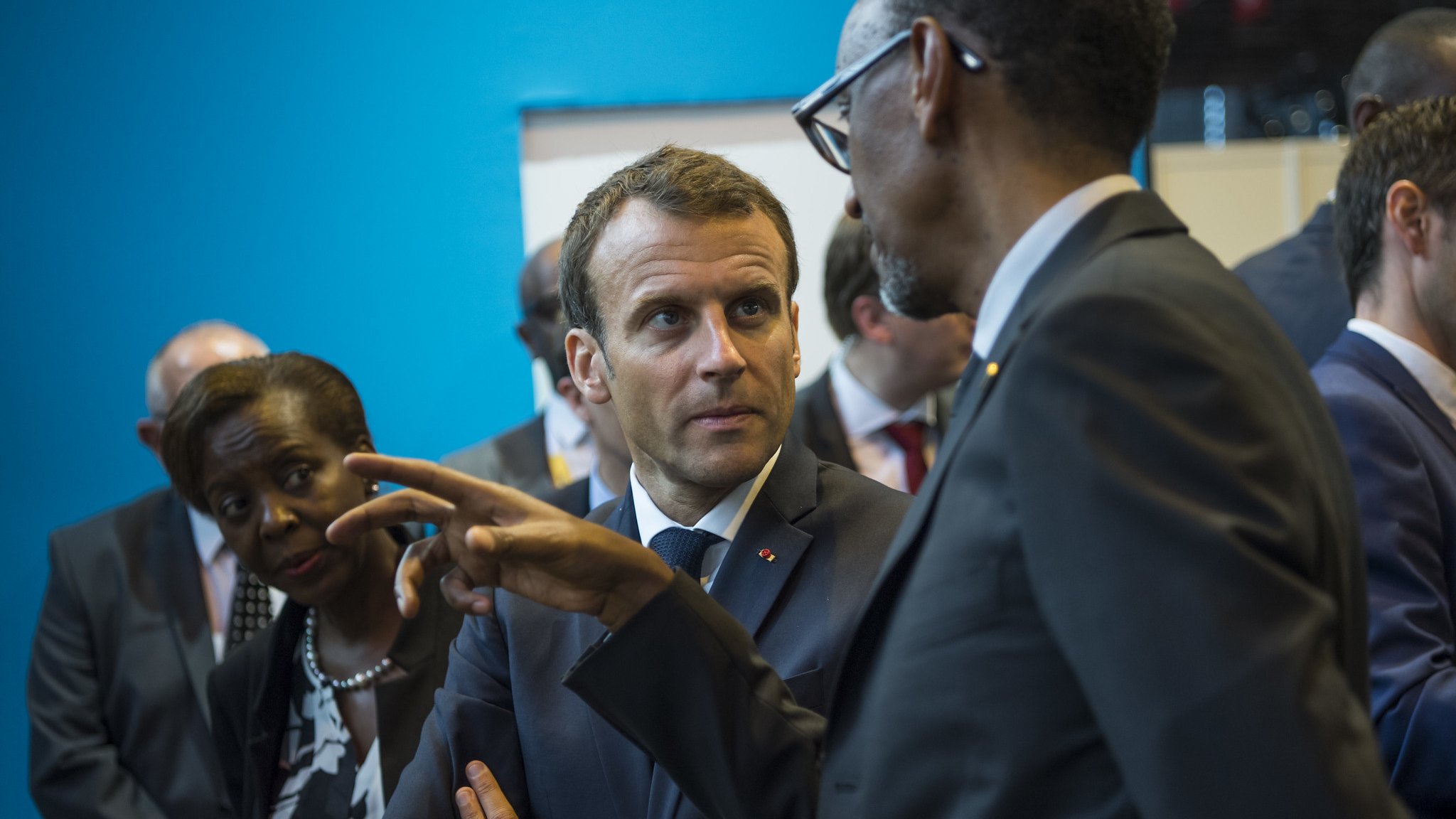 L'image montre un groupe de personnes en conversation dans un environnement qui semble formel. Au premier plan, un homme en costume sombre, probablement un leader, discute avec un autre homme, qui porte également un manteau formel. Le premier homme a une expression attentive, se concentrant sur la discussion, tandis que le second gesticule avec sa main, comme s'il faisait un point important. En arrière-plan, une femme les observe, affichant une expression de sérieux. Le décor est composé de murs d'un bleu vif, ce qui crée un contraste avec les costumes sombres des personnages. L'atmosphère semble professionnelle et engagée, suggérant une rencontre importante.