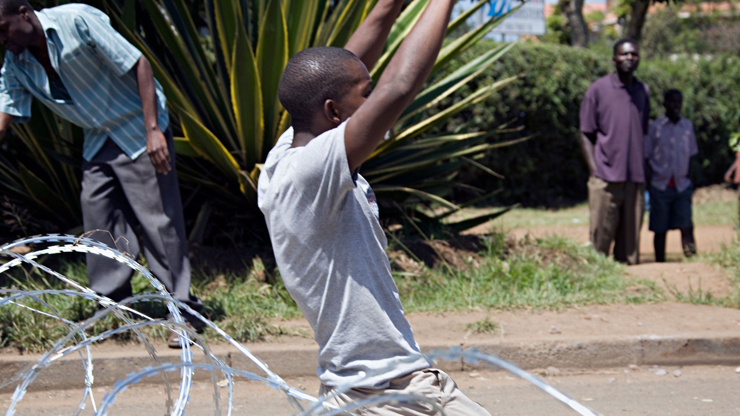 L'image présente une scène animée dans un environnement urbain. Au premier plan, un jeune homme est agenouillé sur le sol, les bras levés dans une posture d'expression intense, peut-être de joie ou de victoire. Il porte un t-shirt gris et un pantalon clair. Autour de lui, des fil de fer barbelés sont dispersés, ajoutant une dimension de tension à la scène. En arrière-plan, plusieurs personnes sont visibles, certaines observant, d'autres engagées dans des activités, suggérant une atmosphère collective. La végétation, avec des plantes vertes, donne un aspect naturel au lieu. L'ensemble de cette image transmet une forte émotion, où les gestes et les postures des individus racontent une histoire d'énergie et de dynamisme.