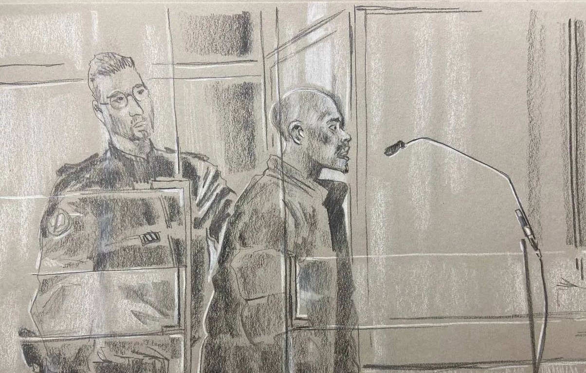 L'image représente une scène d'audience judiciaire. Au premier plan, on voit un homme debout, probablement un accusé, qui se tient derrière un panneau de séparation, exprimant une attitude sérieuse. Il est entouré par des éléments du tribunal, comme un microphone devant lui, indiquant qu'il pourrait s'exprimer. À sa gauche se trouve un policier en uniforme, affichant une posture attentive et vigilant. L'atmosphère de l'image est solennelle, reflétant le sérieux de la situation judiciaire. Les couleurs utilisées sont principalement des nuances de gris, et le style est esquissé, avec un rendu qui transmet les émotions et la tension de l'instant.