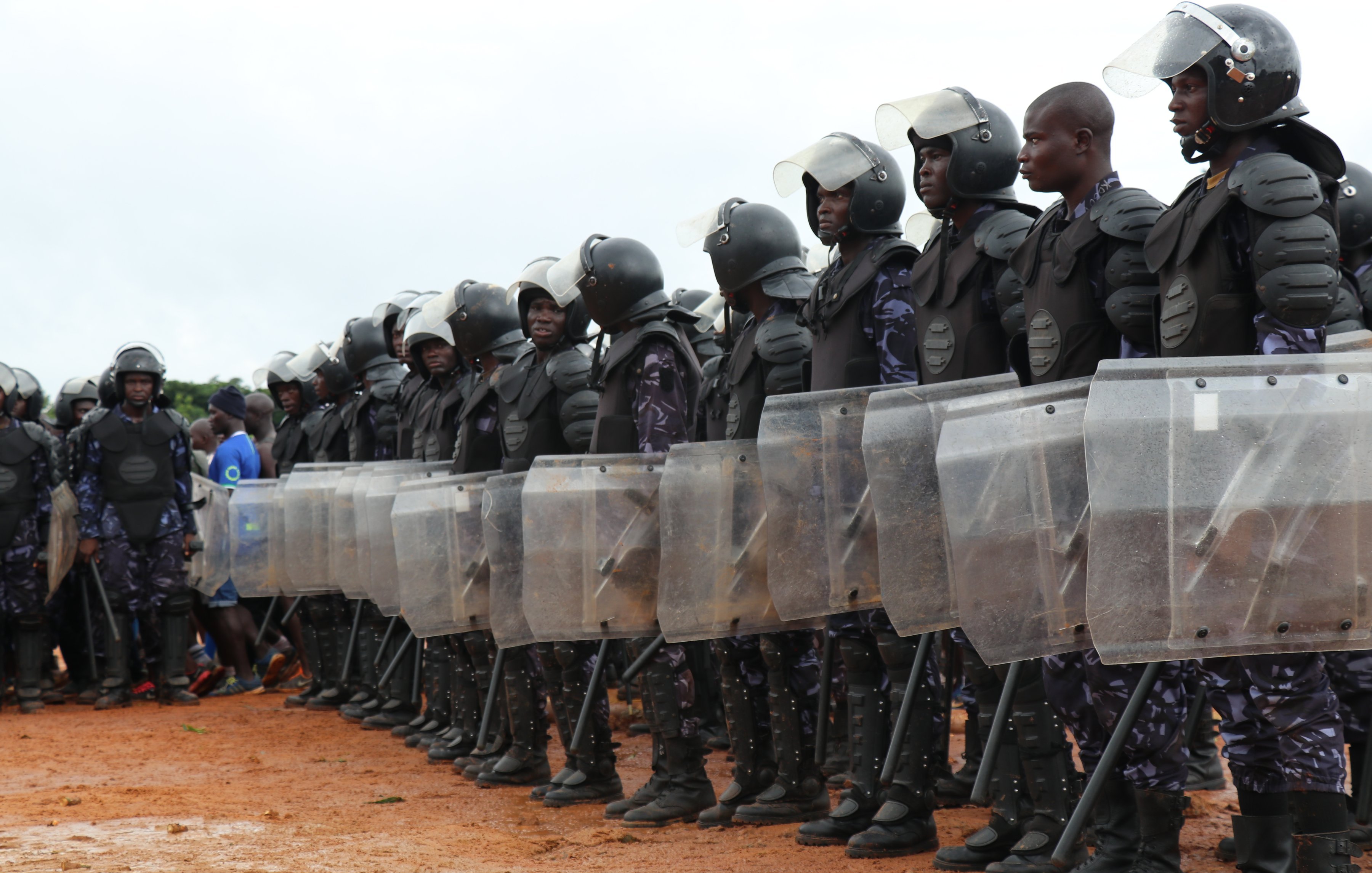L'image montre une formation de policiers en tenue anti-émeute. Ils se tiennent en ligne, rigides et organisés, en portant des casques de protection et des gilets pare-balles. Chaque policier tient un bouclier transparent en face de lui, prêt à faire face à une situation potentielle. Le sol est en terre battue et l'environnement semble être à l'extérieur, avec quelques nuages visibles dans le ciel. Au loin, on peut apercevoir des personnes qui observaient la scène. L'atmosphère suggère une présence de sécurité renforcée.