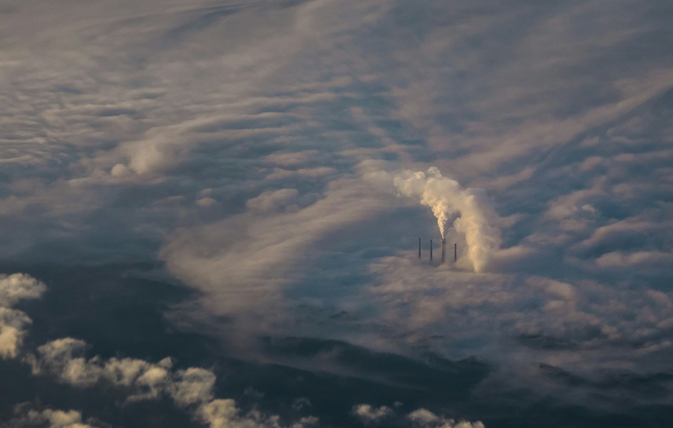 L'image présente une vue aérienne d'un paysage nuageux. On peut distinguer plusieurs cheminées d'une centrale, émettant des colonnes de fumée blanche qui se fondent dans les nuages environnants. Les nuages eux-mêmes paraissent denses et vaporeux, créant une atmosphère mystique. La lumière semble filtrer à travers le brouillard, ajoutant une touche douce au paysage. Dans l'ensemble, l'image évoque une impression de grandeur et de puissance, avec la nature et l'industrialisation coexistant dans un environnement nuageux.