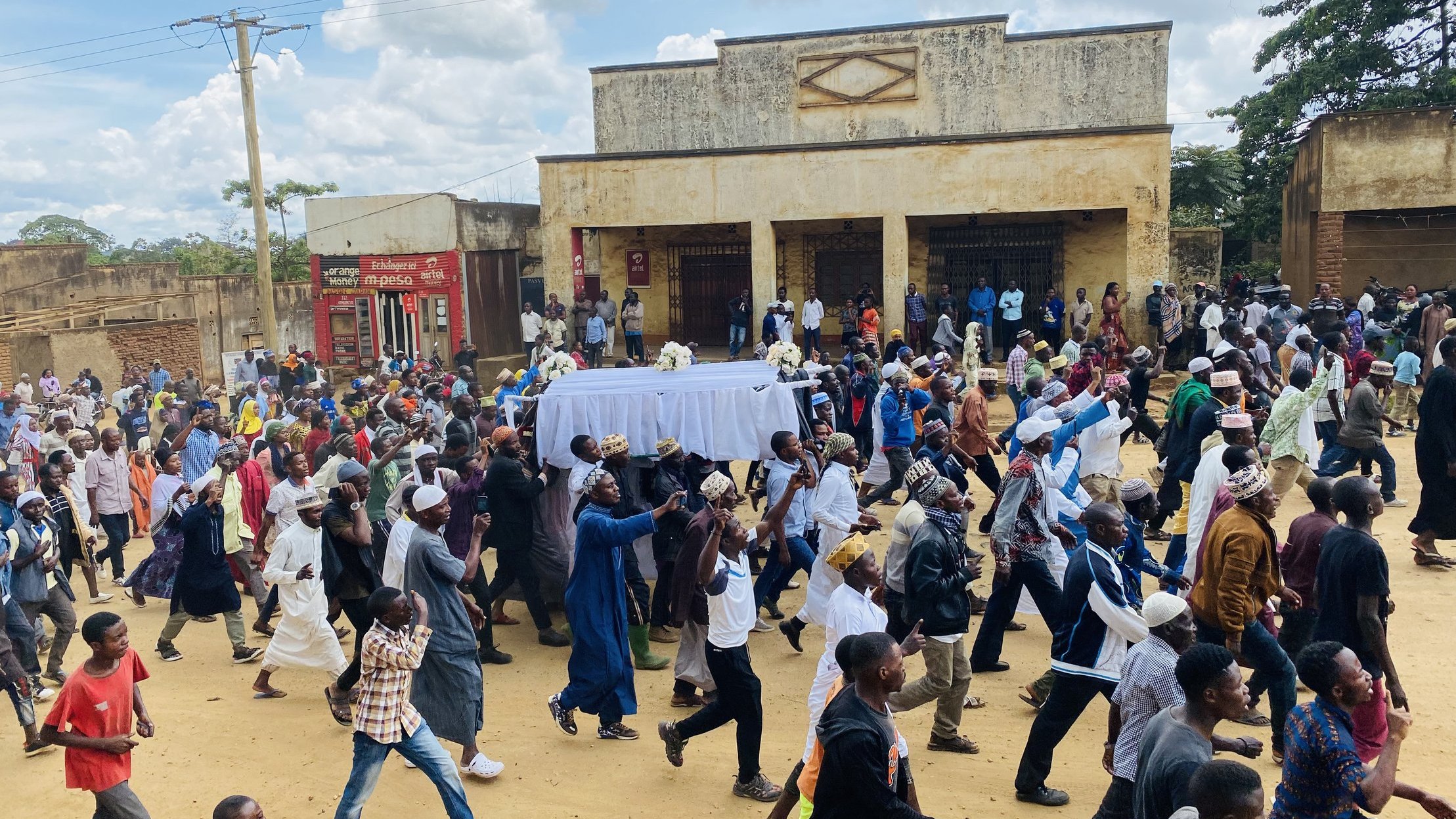 L'image montre une scène animée dans une rue où une grande foule se déplace. Au centre de l'attention, des personnes portent un drap blanc qui semble recouvrir un cercueil, indiquant qu'il s'agit d'un cortège funéraire. Les membres de la foule sont vêtus de divers habits, certains en tenues traditionnelles, d'autres plus contemporaines. Les visages reflètent des émotions variées, allant du recueillement à la tristesse. À l'arrière-plan, on peut remarquer des bâtiments aux murs fissurés, avec des panneaux publicitaires visibles. Le ciel est légèrement nuageux, créant une atmosphère à la fois sombre et lugubre, mais la scène reste vibrante de vie grâce à la présence de nombreux participants.