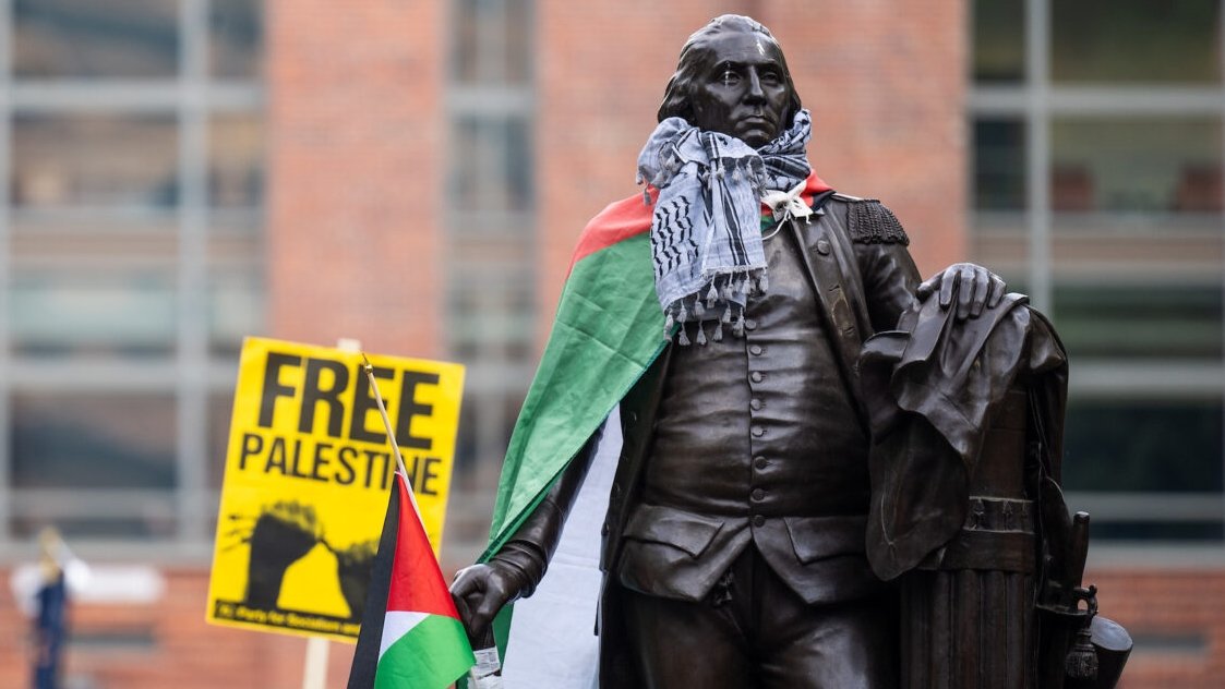 L'image montre une statue en bronze d'un homme debout, tenant un bâton dans une main et un document dans l'autre. Cette statue est habillée d'un keffieh, un foulard traditionnel palestinien en noir et blanc, et elle est drapée d'un drapeau palestinien au motif rouge, blanc, noir et vert. En arrière-plan, on aperçoit un panneau jaune sur lequel est inscrit "FREE PALESTINE" en lettres noires, exprimant un message de soutien pour la cause palestinienne. L'environnement semble être une place publique animée, avec des bâtiments en briques visibles derrière. Cette scène témoigne de la mobilisation et de la solidarité, créant une ambiance engageante et militante.