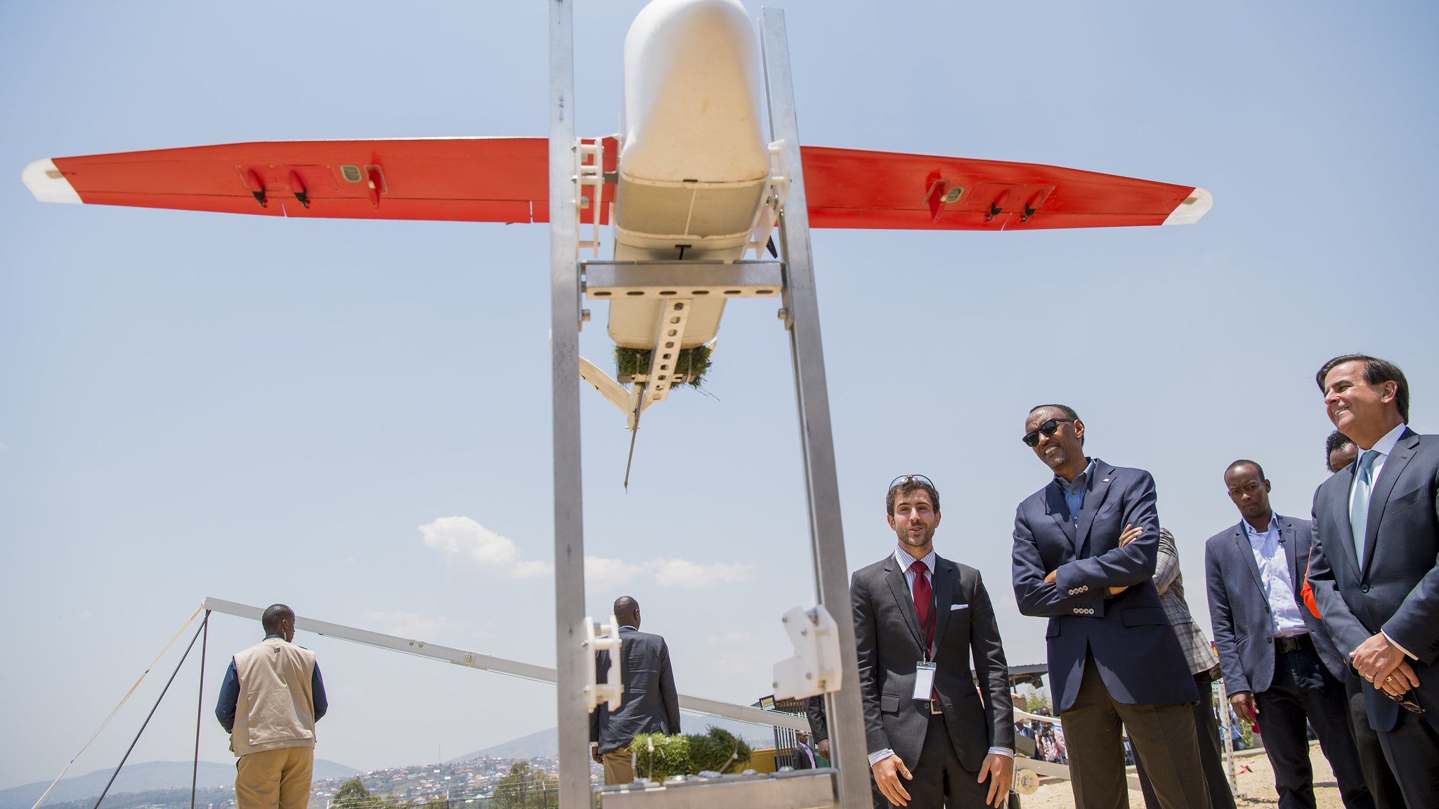 L'image montre un drone de forme allongée, aux ailes rouges et au corps blanc, placé sur une structure métallique. Plusieurs personnes, notamment des hommes en costumes, se tiennent à proximité, observant le drone avec intérêt. En arrière-plan, on aperçoit un paysage urbain et des collines sous un ciel bleu lumineux. L'atmosphère semble à la fois sérieuse et enthousiaste, suggérant un événement lié à la technologie ou à l'innovation.